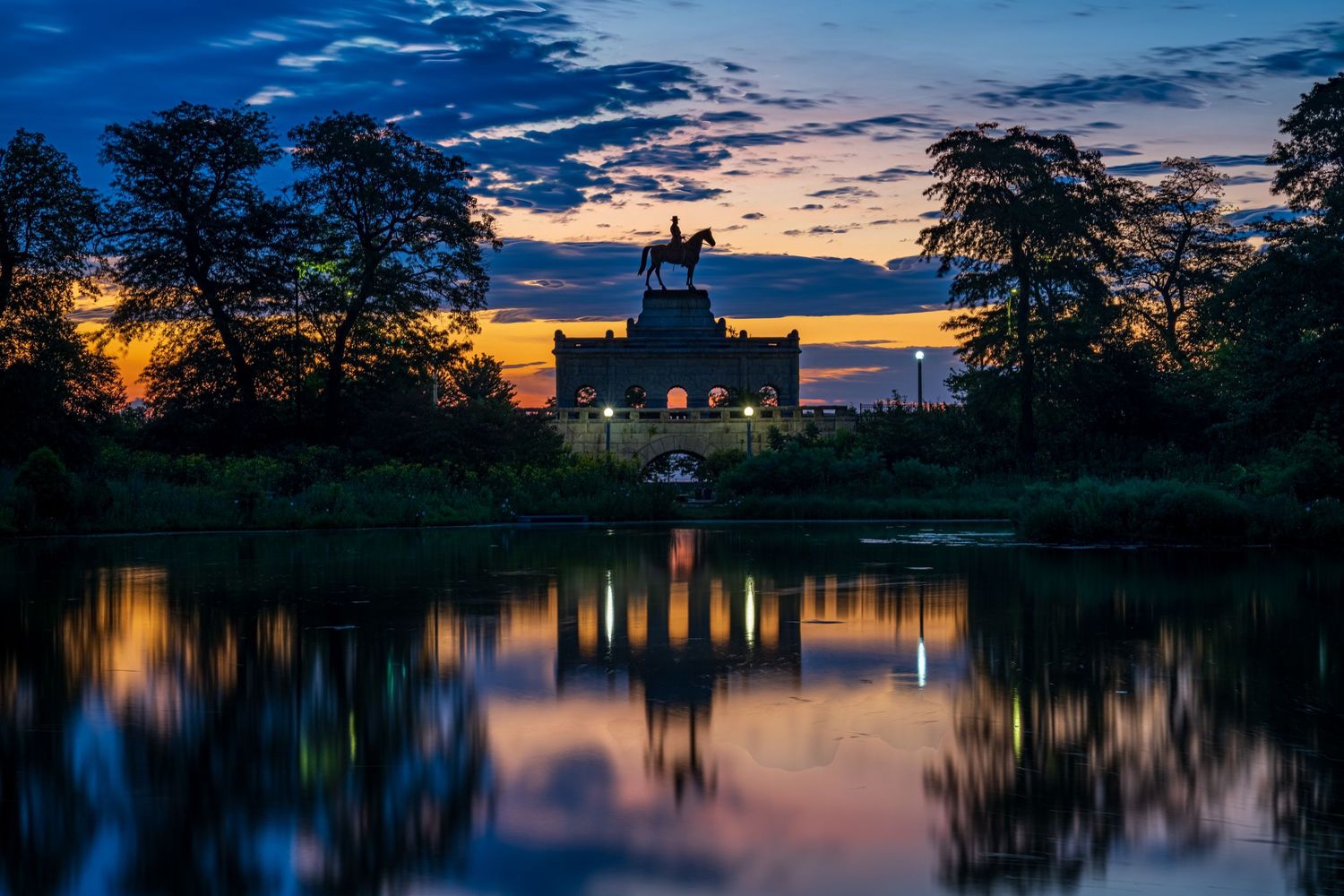Twilight reflection of an equestrian statue and castle silhouetted against a vibrant sunset sky mirrored in a calm lake.