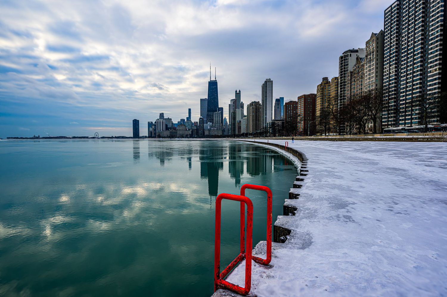 Chicago skyline reflects on Lake Michigan in winter, with snow-covered shore and red barriers in the foreground.
