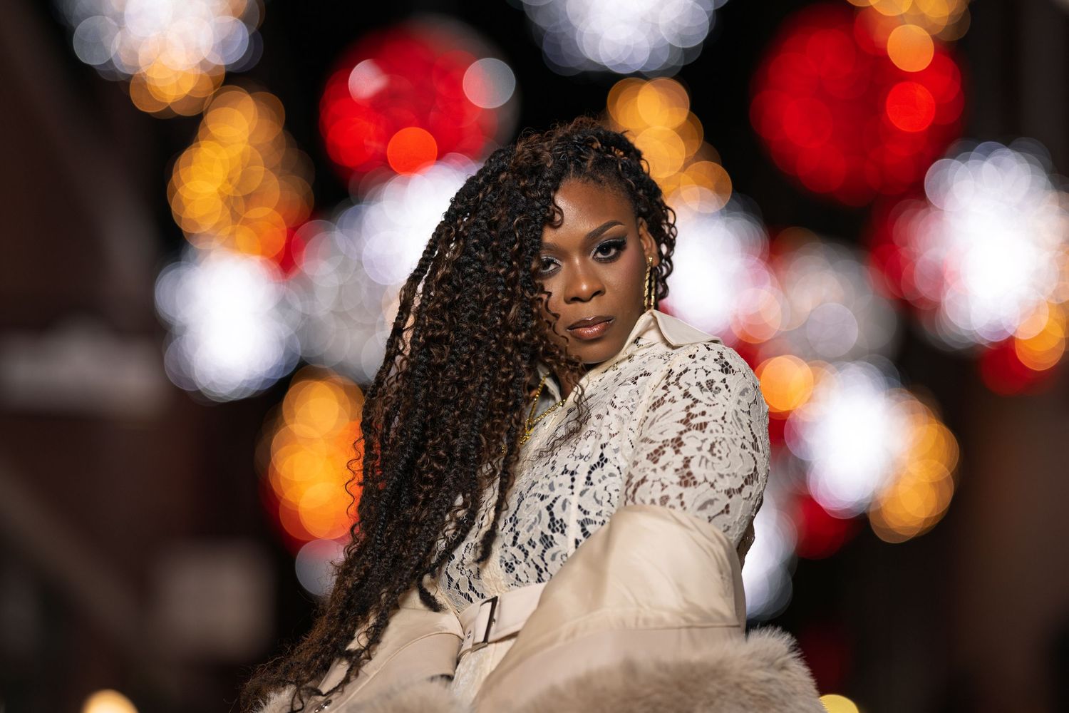 A young Black woman with long braided hair poses in a lace jacket against festive bokeh Christmas lights.