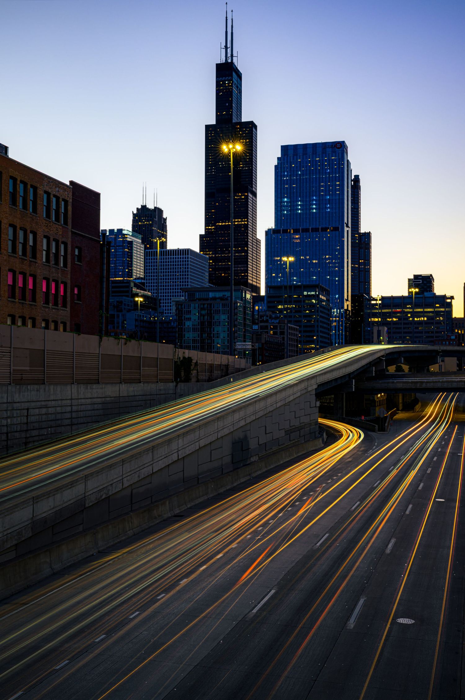 Long exposure shot of Chicago highway light trails at dusk with Willis Tower and city skyline in background.