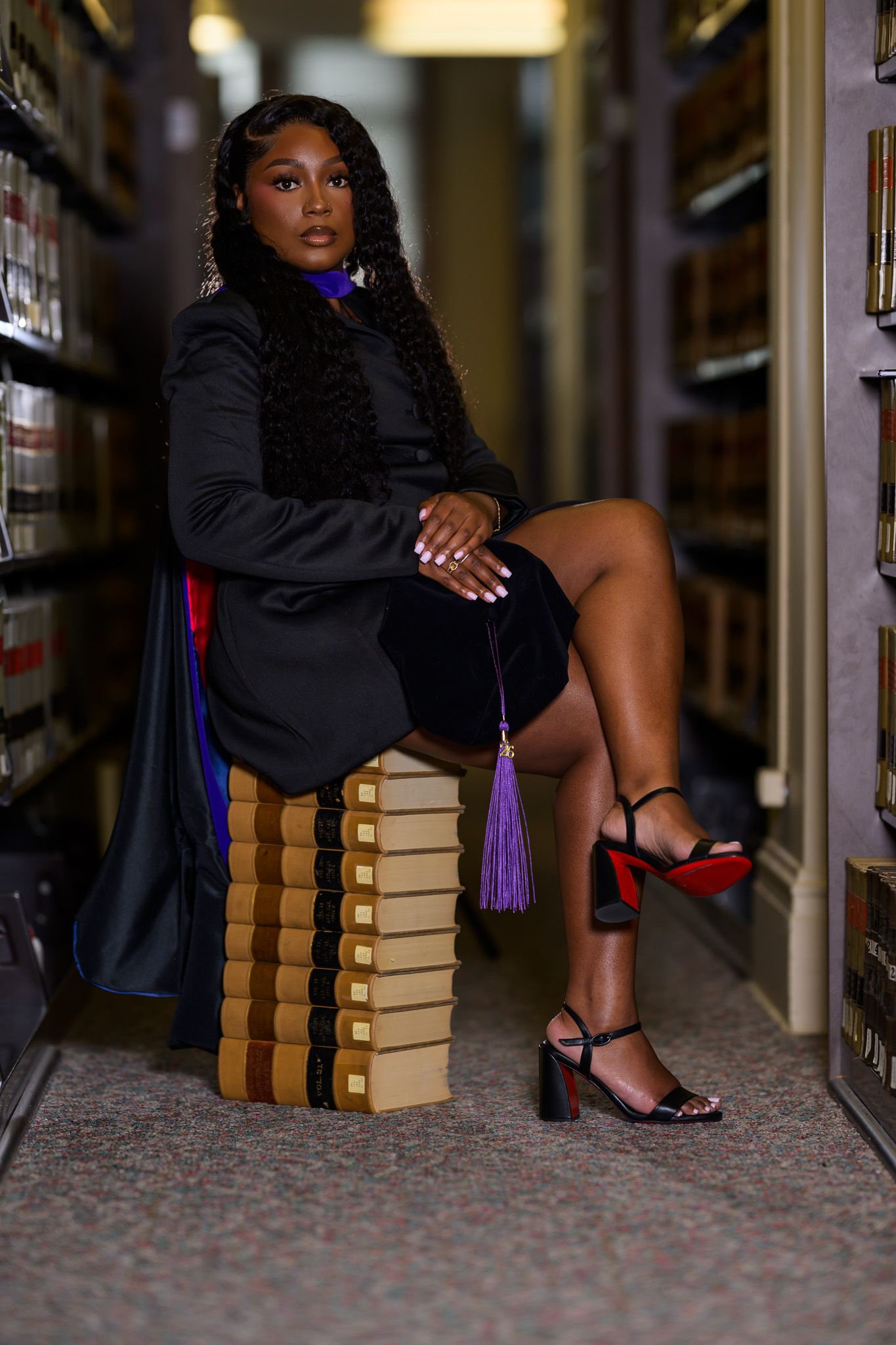 A graduating woman in black academic regalia sits on stacked books in a library aisle, holding a purple tassel.