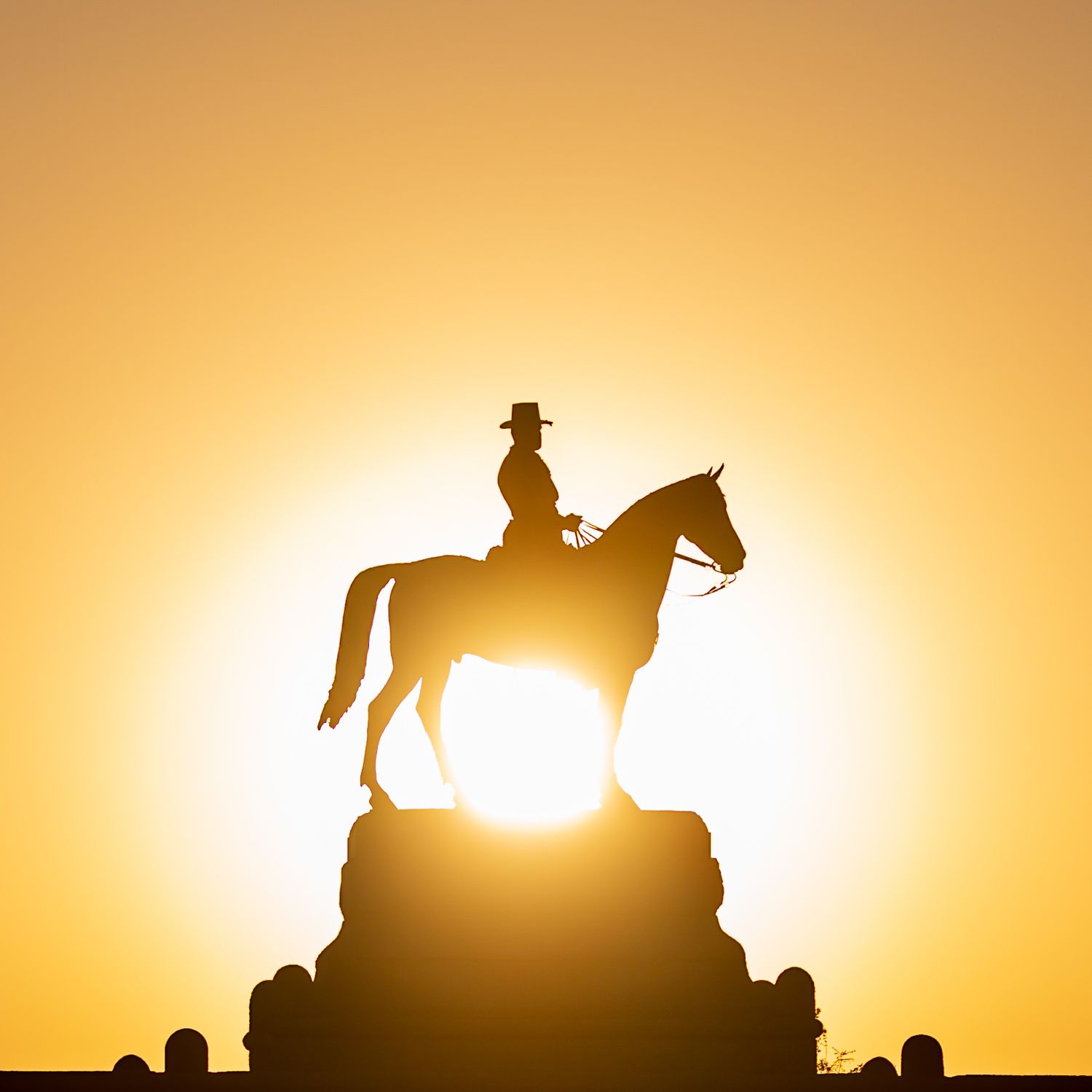Silhouette of an equestrian statue with a rider in a cowboy hat against a golden sunset sky.