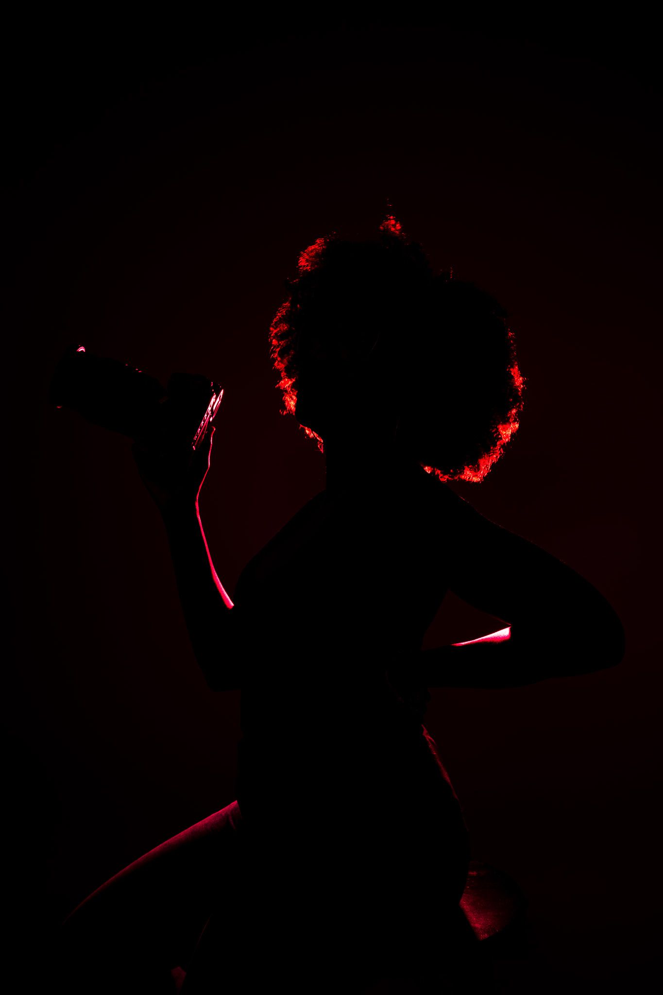 Silhouette of a person with curly hair holding a microphone, illuminated by dramatic red backlighting.