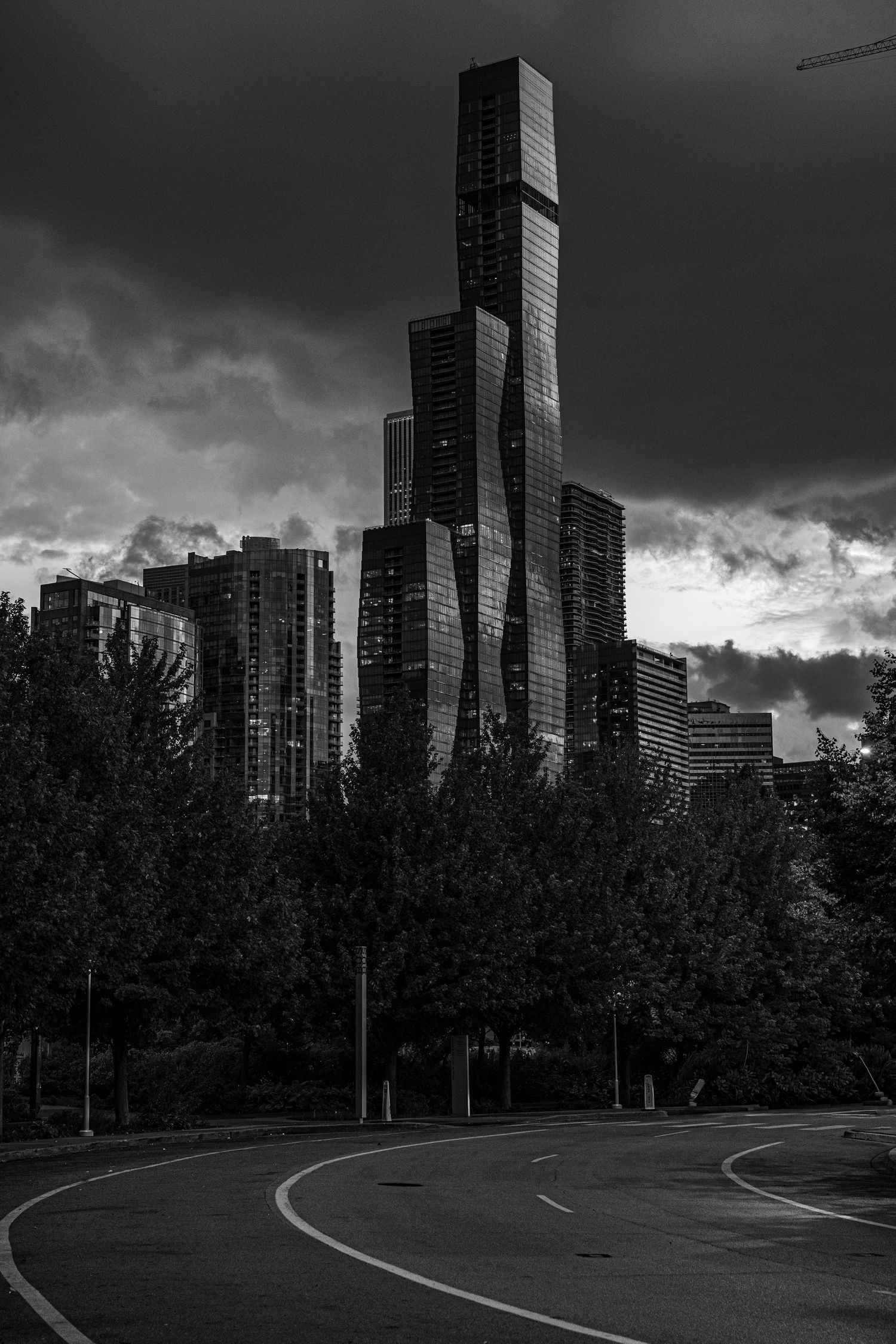 Black and white photo of a dramatic skyscraper skyline under stormy clouds with trees and a curved road in the foreground.