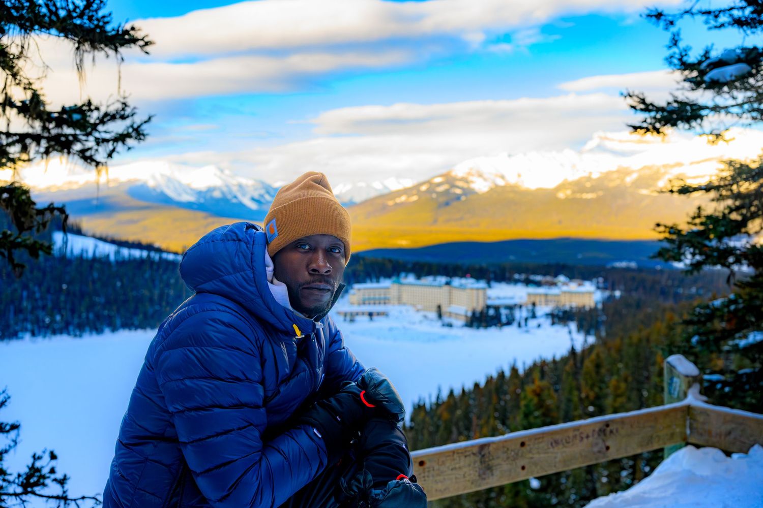 Man in blue jacket and orange beanie stands on snowy overlook with mountain resort and golden sunset sky.
