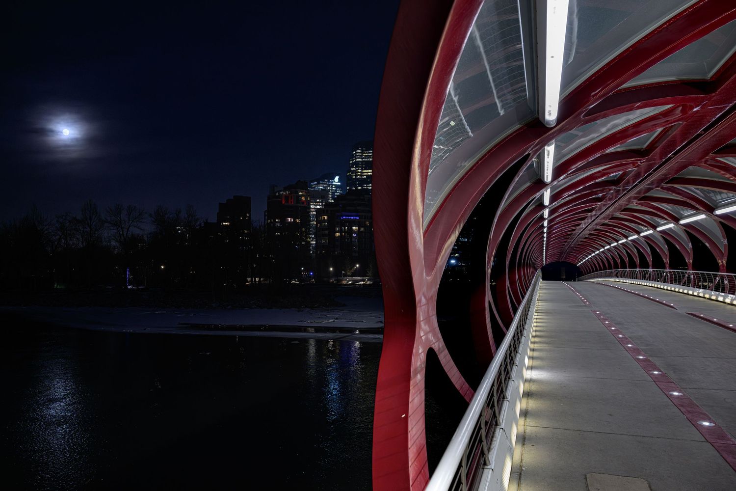 Red arched pedestrian bridge at night with illuminated walkway and city skyline reflected on water under a full moon.