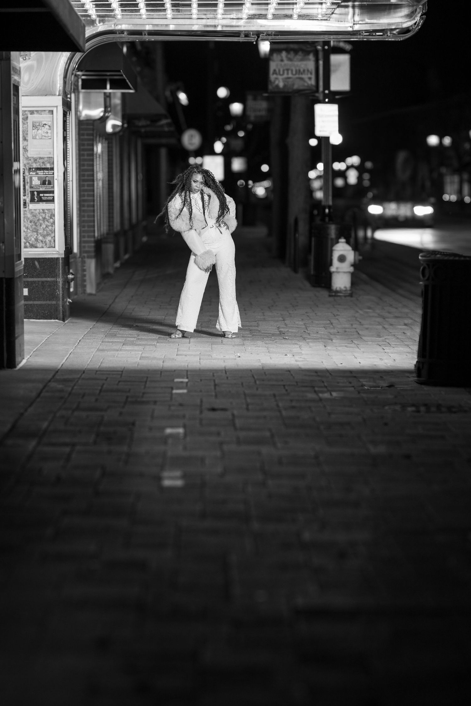 Black and white night street photo of a woman in wide-leg pants posing on a city sidewalk.