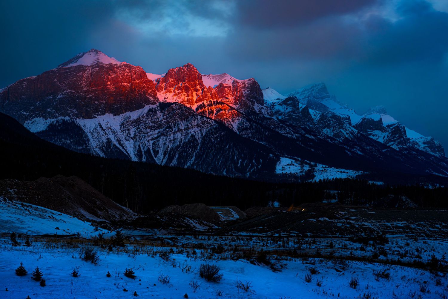 Snow-covered mountain peaks glowing red at sunset under dramatic stormy skies in a winter alpine landscape.