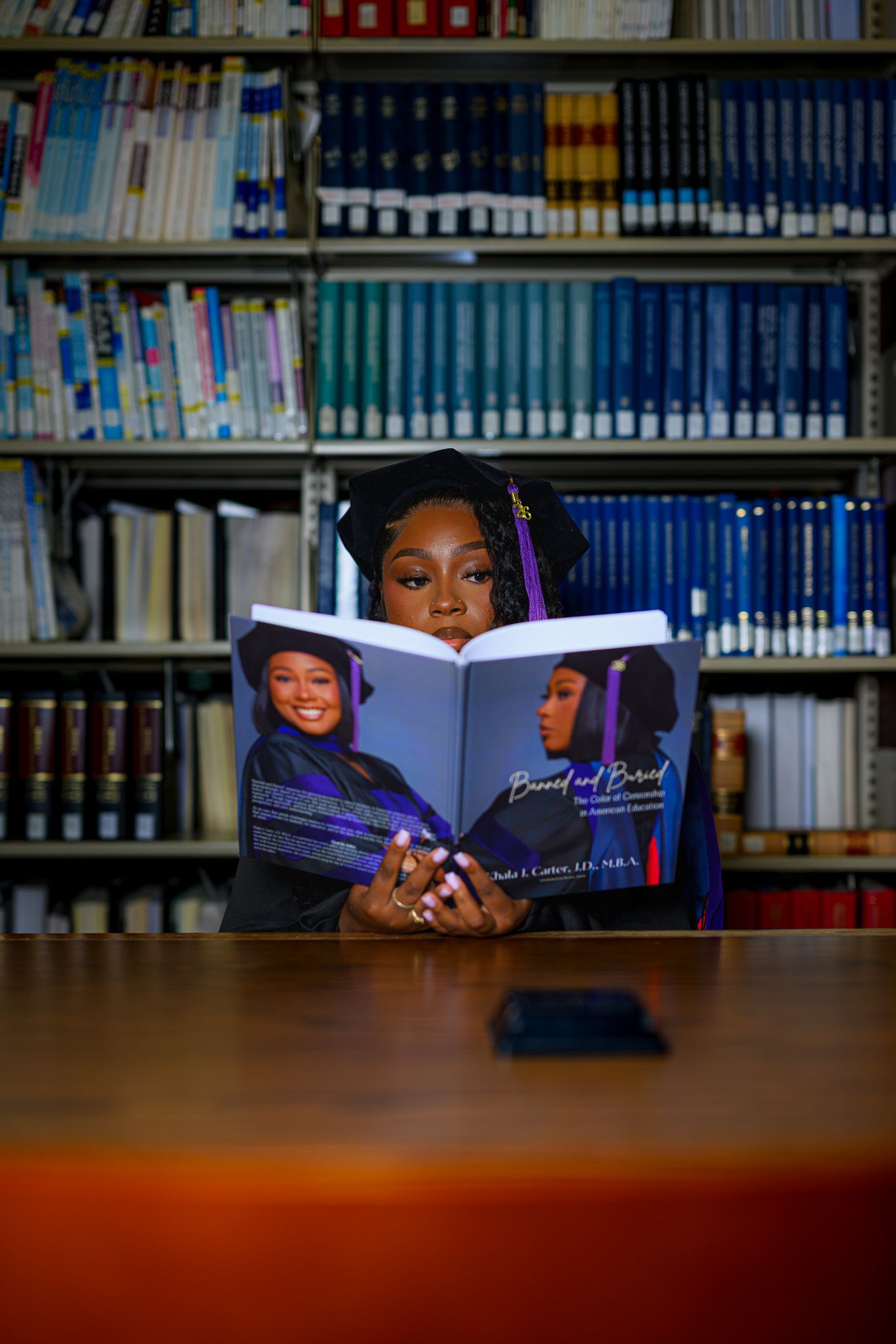Graduate in cap and gown reads her own book in a library, showcasing academic achievement and authorship.