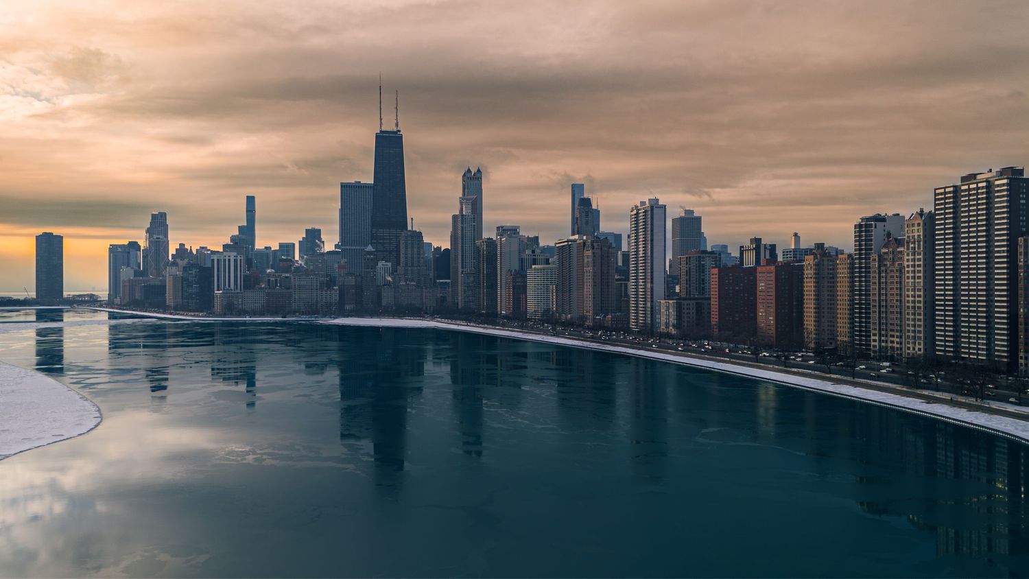 Chicago skyline reflecting on calm Lake Michigan waters at sunset with dramatic cloudy sky.
