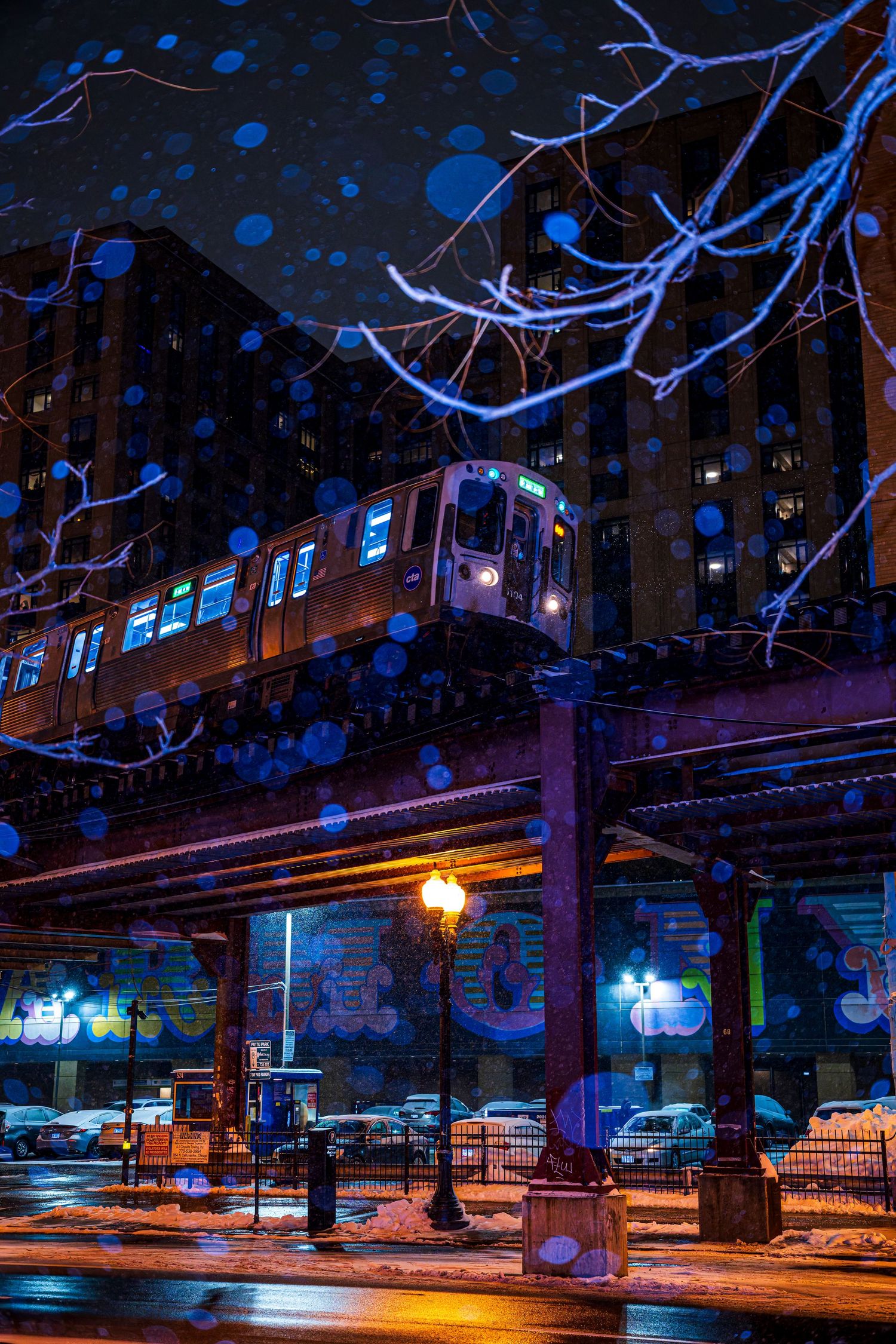 Chicago elevated train passes over a snowy urban street at night, with blue bokeh snowflakes and glowing city lights below.