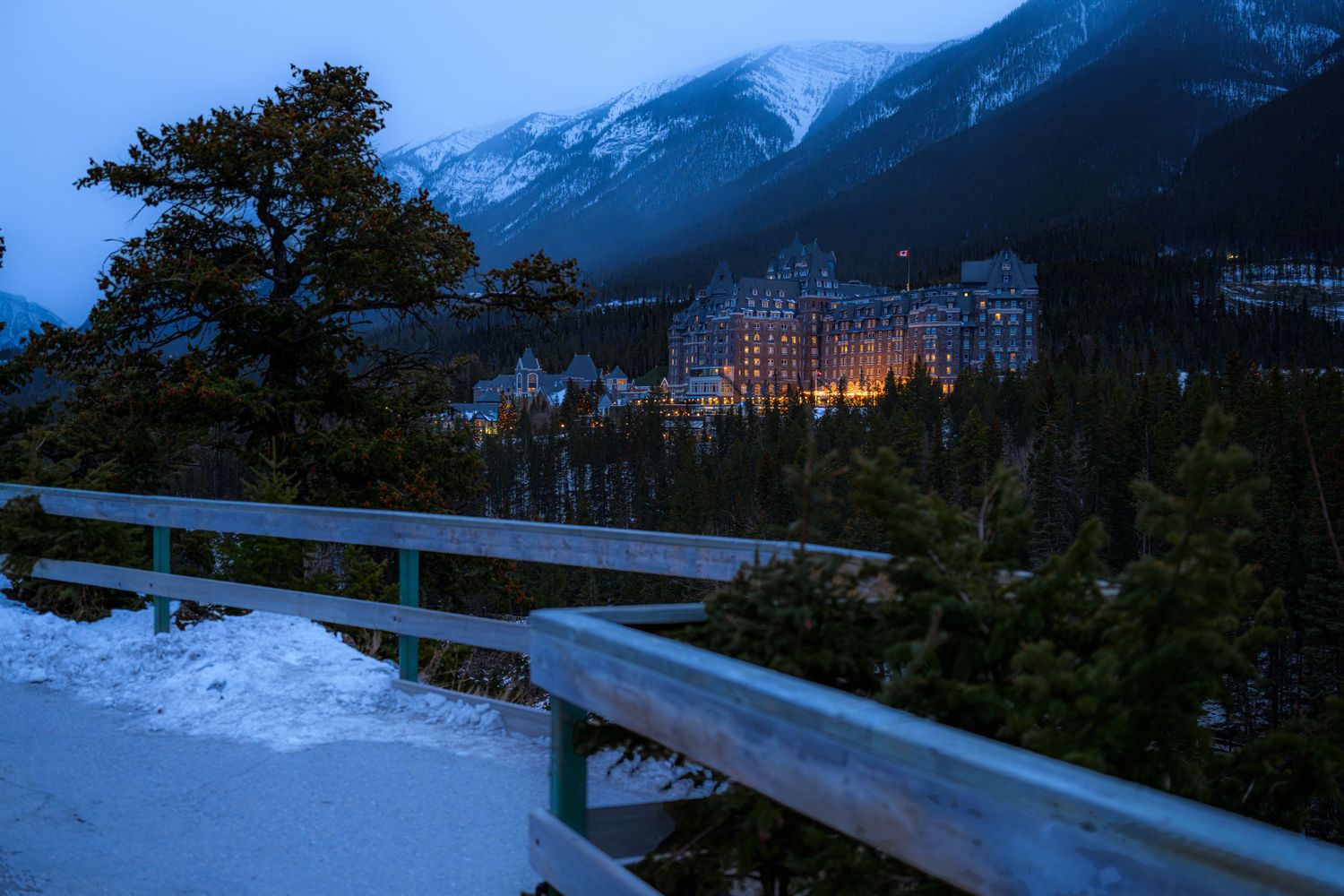 Fairmont Banff Springs Hotel glows at dusk surrounded by snow-dusted pine trees and Rocky Mountains in Banff, Canada.