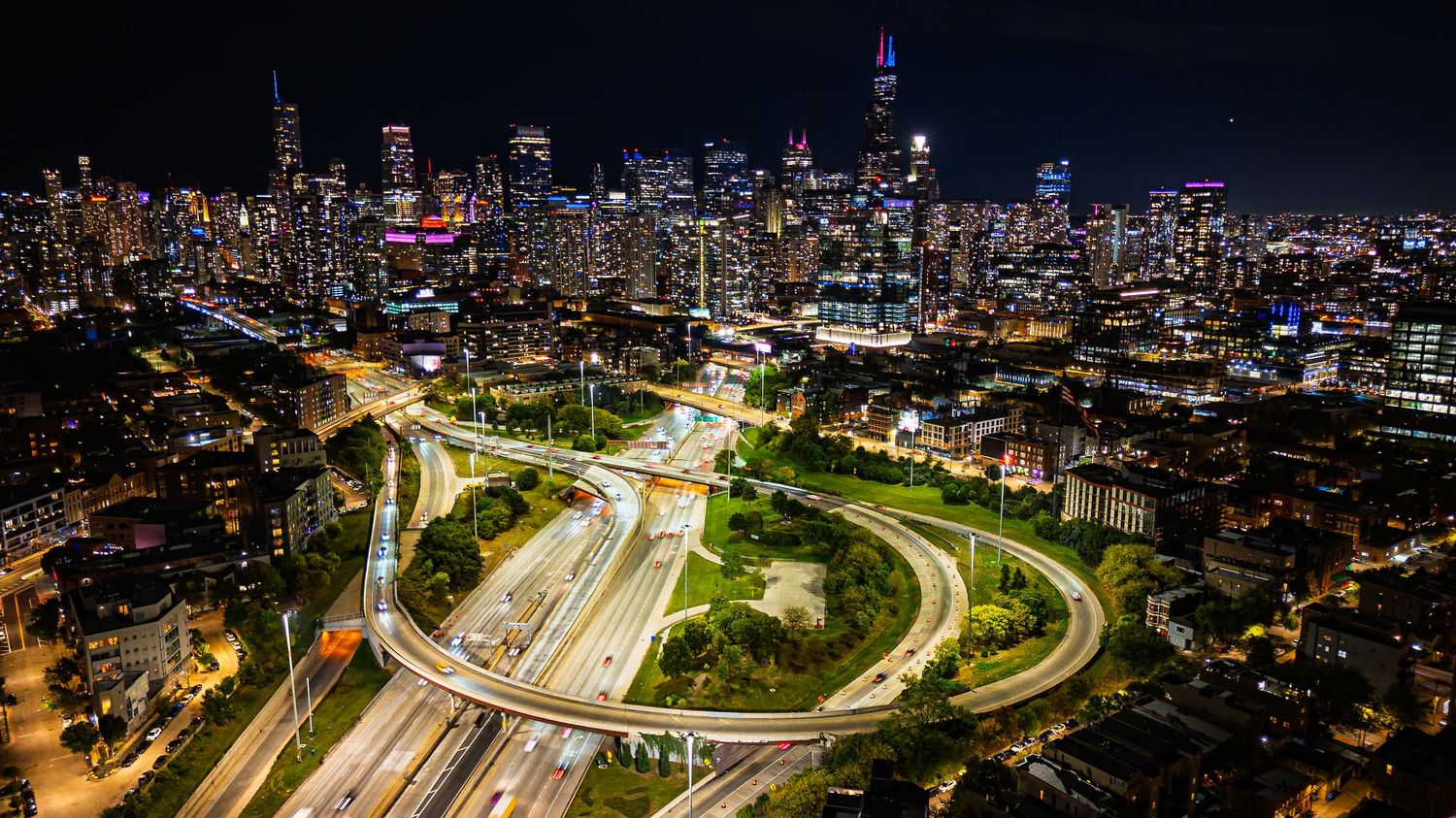 Aerial night view of a glowing city skyline with illuminated highway interchange and light trails from traffic.