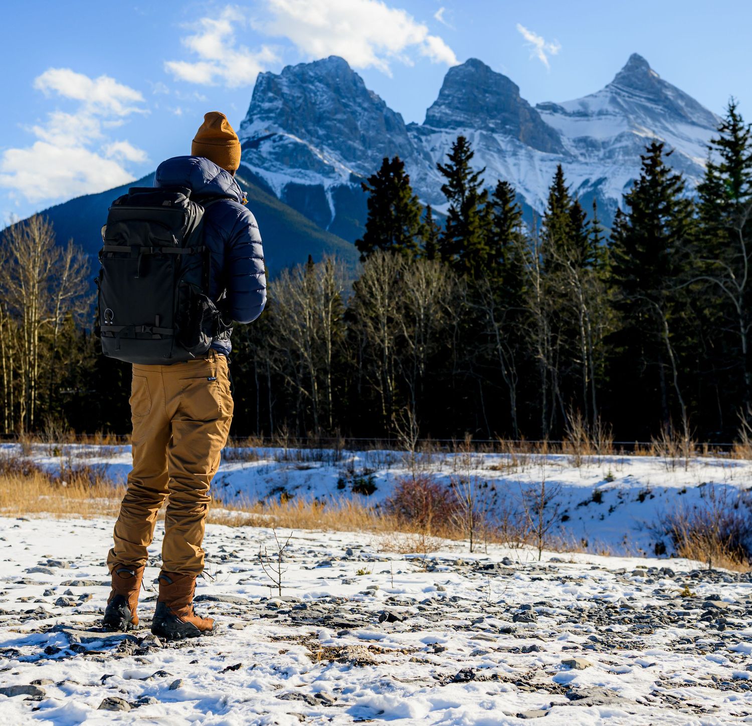 Hiker with backpack and yellow beanie standing in snowy field gazing at Three Sisters mountains in Canmore, Alberta.