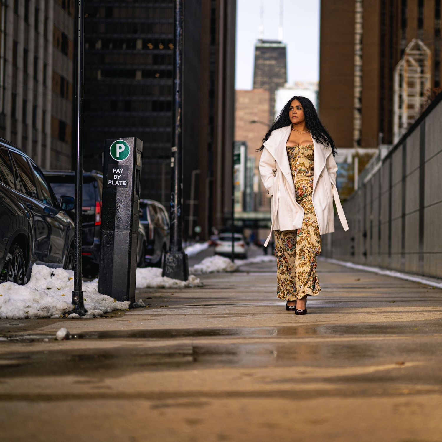 Woman in gold sequin dress and white coat walks confidently on a wet urban sidewalk with city buildings and snow.