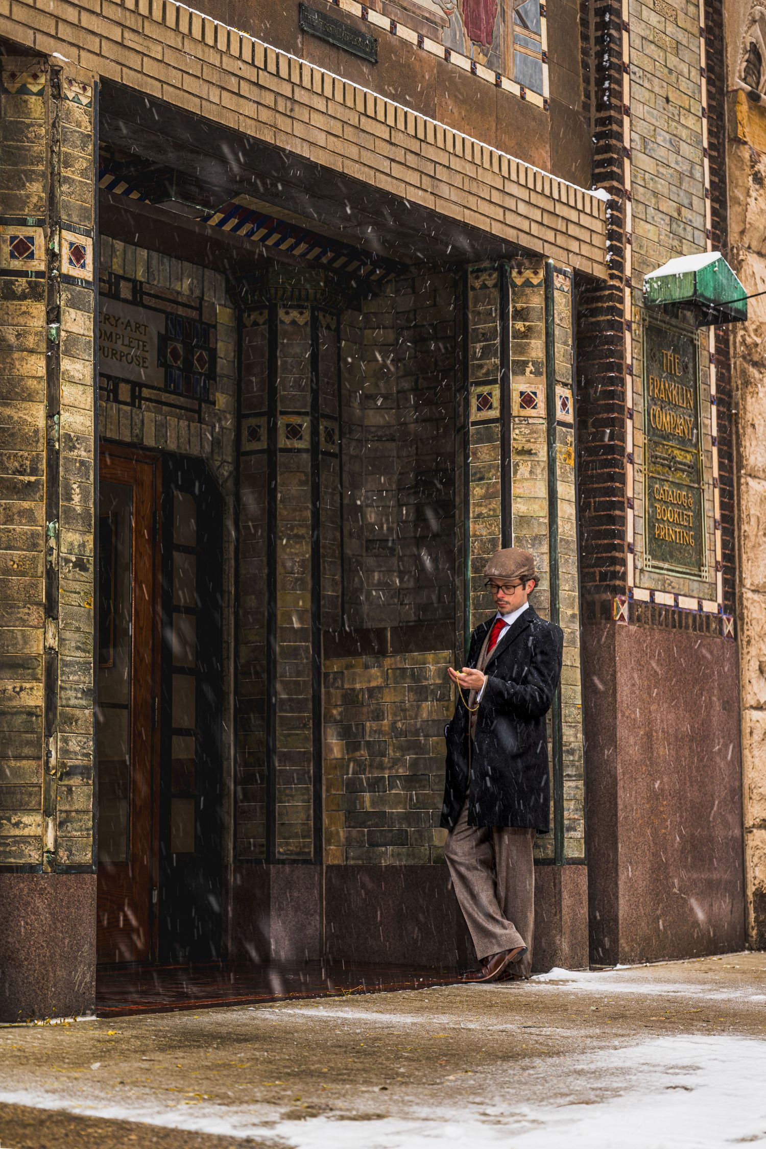 Man in hat and black coat checks phone outside ornate brick building entrance during light snowfall.
