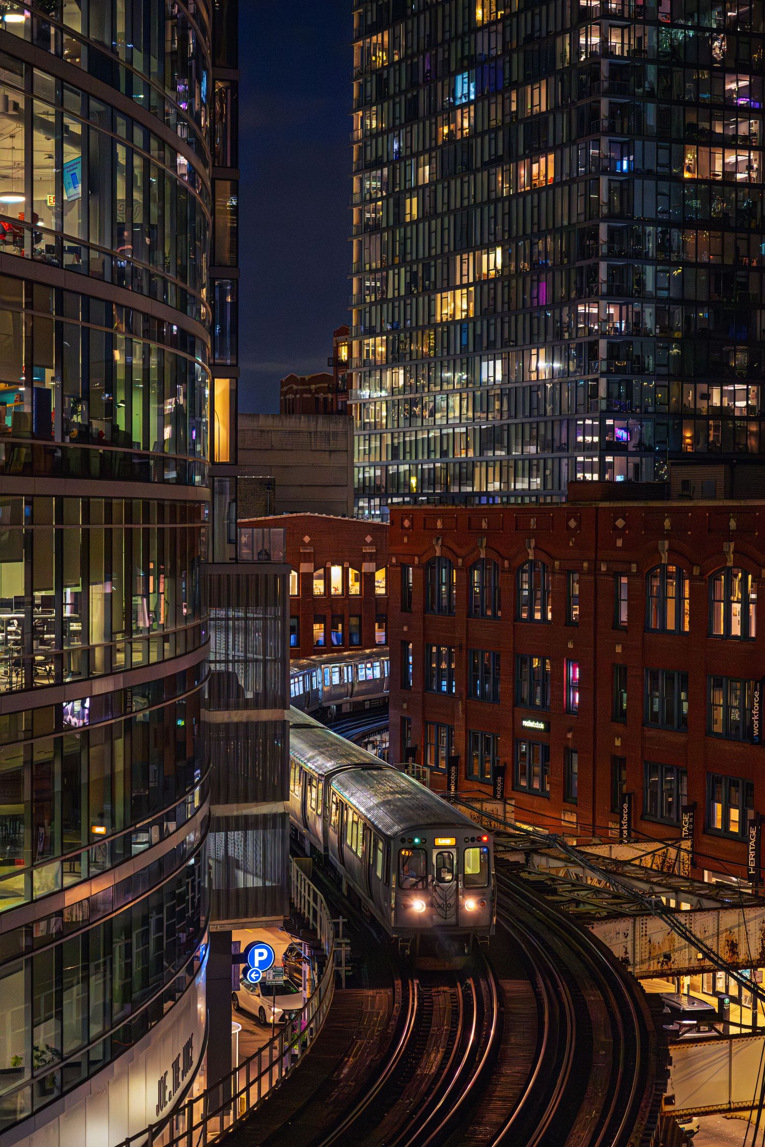 Chicago elevated train curves through illuminated downtown skyscrapers at night, with glowing office buildings and city lights.