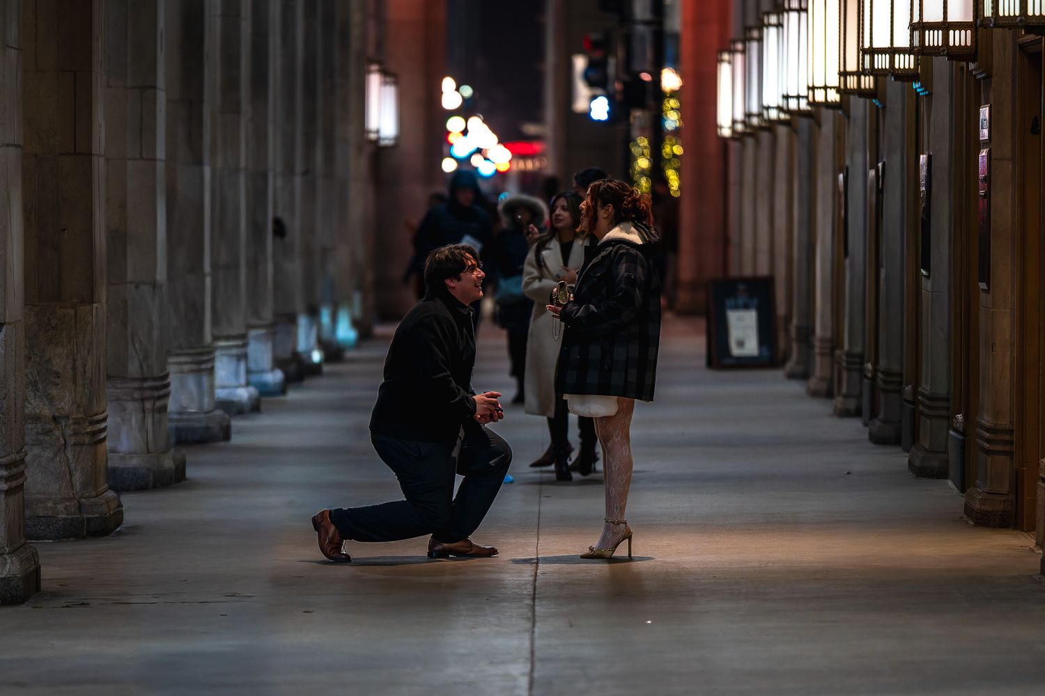 Man proposing to woman on one knee in an illuminated city arcade at night.