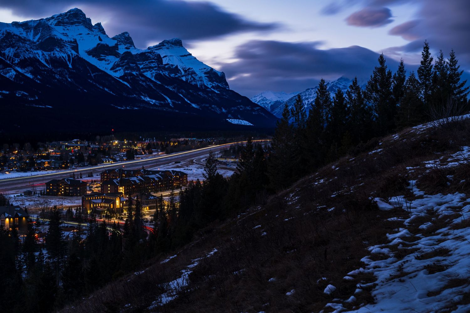 Twilight view of Canmore Alberta with glowing city lights nestled between snow-dusted Rocky Mountains and evergreen forests.