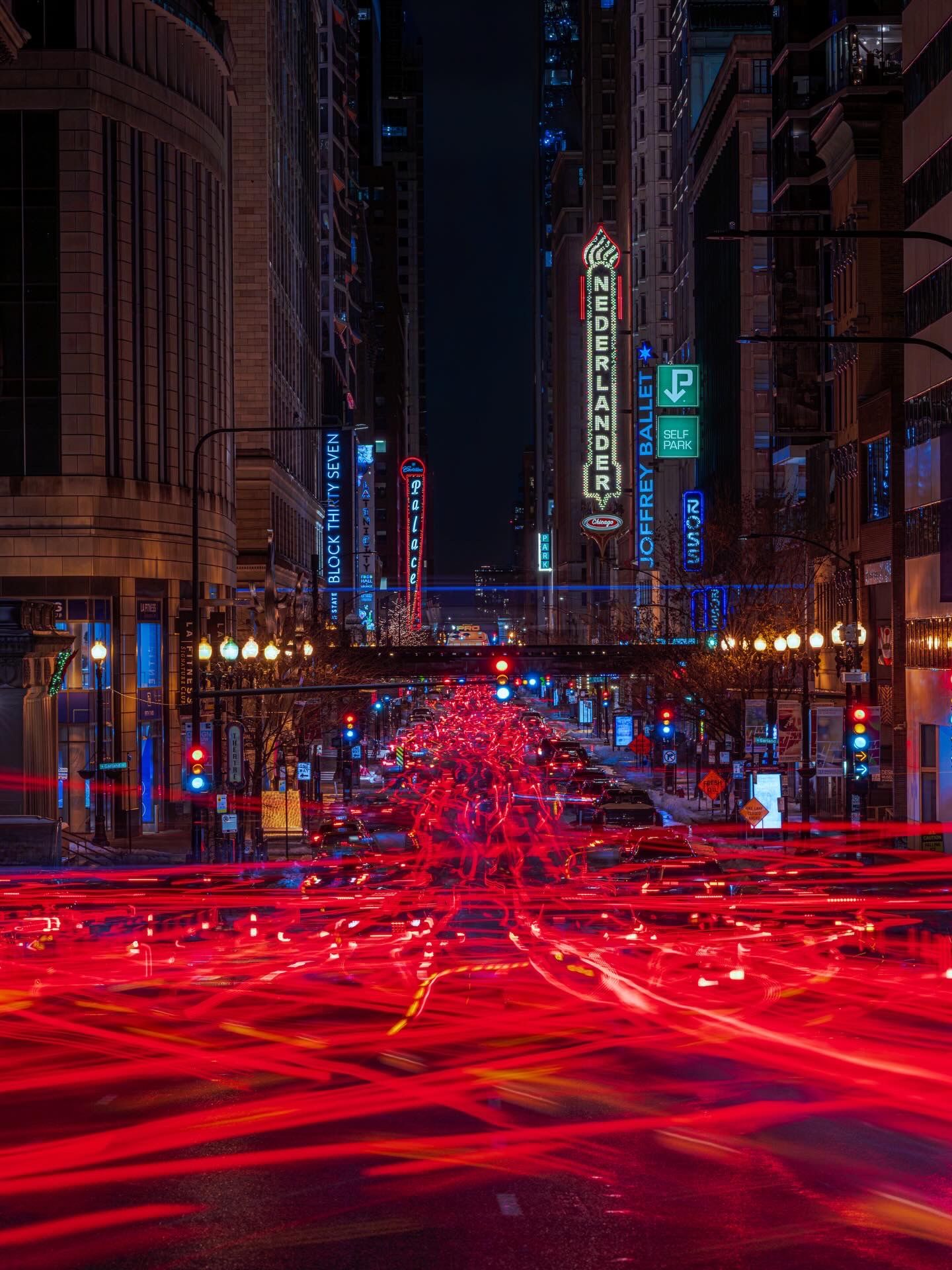 Long-exposure night shot of a busy city street with red light trails, neon signs, and tall buildings lining the road.
