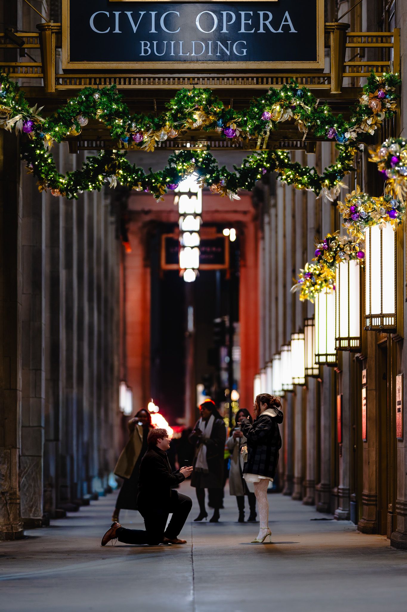 Man proposing to woman in festive Civic Opera Building arcade decorated with Christmas garland lights.
