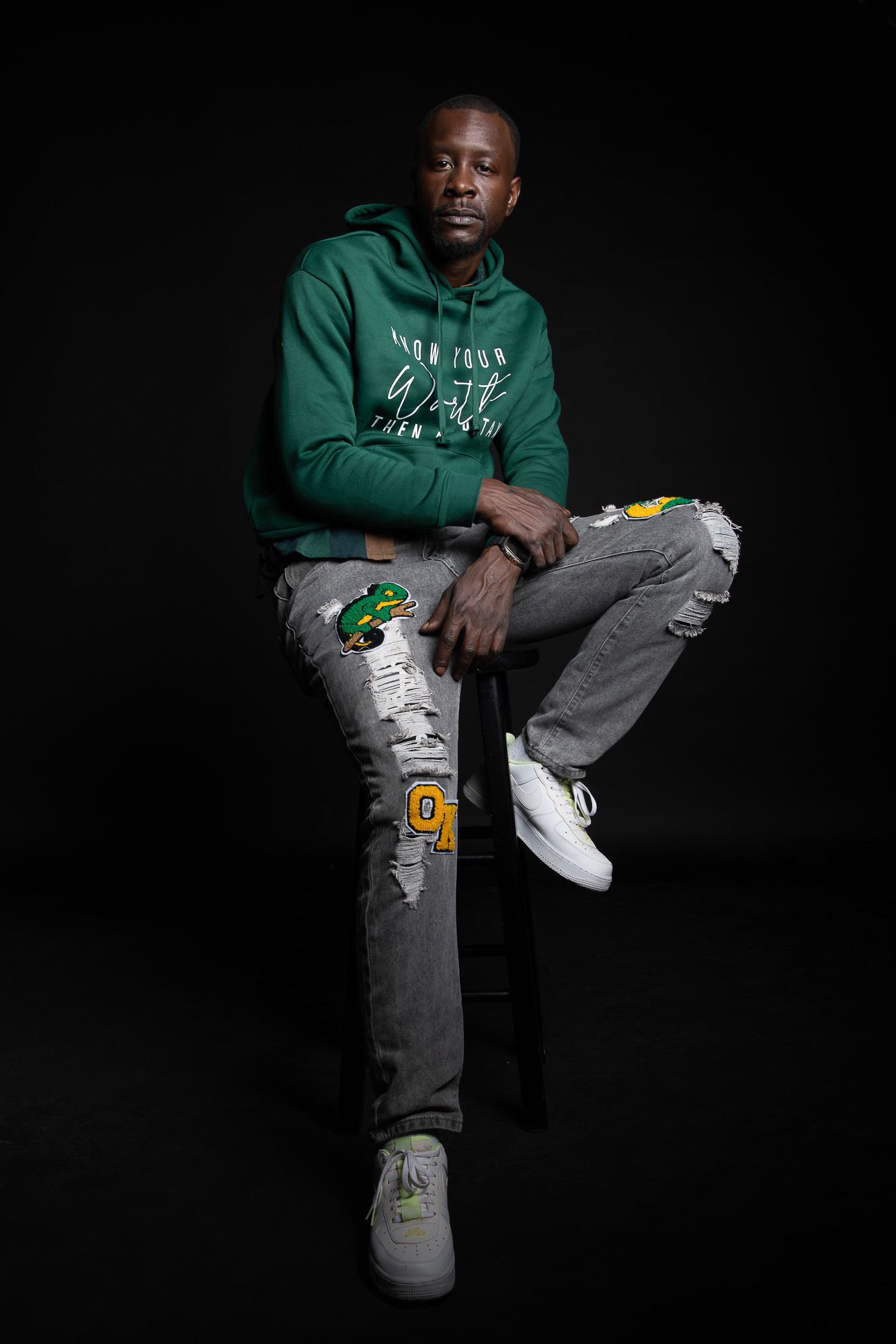 Man in green hoodie and ripped jeans with patches sits confidently on stool against dark background.