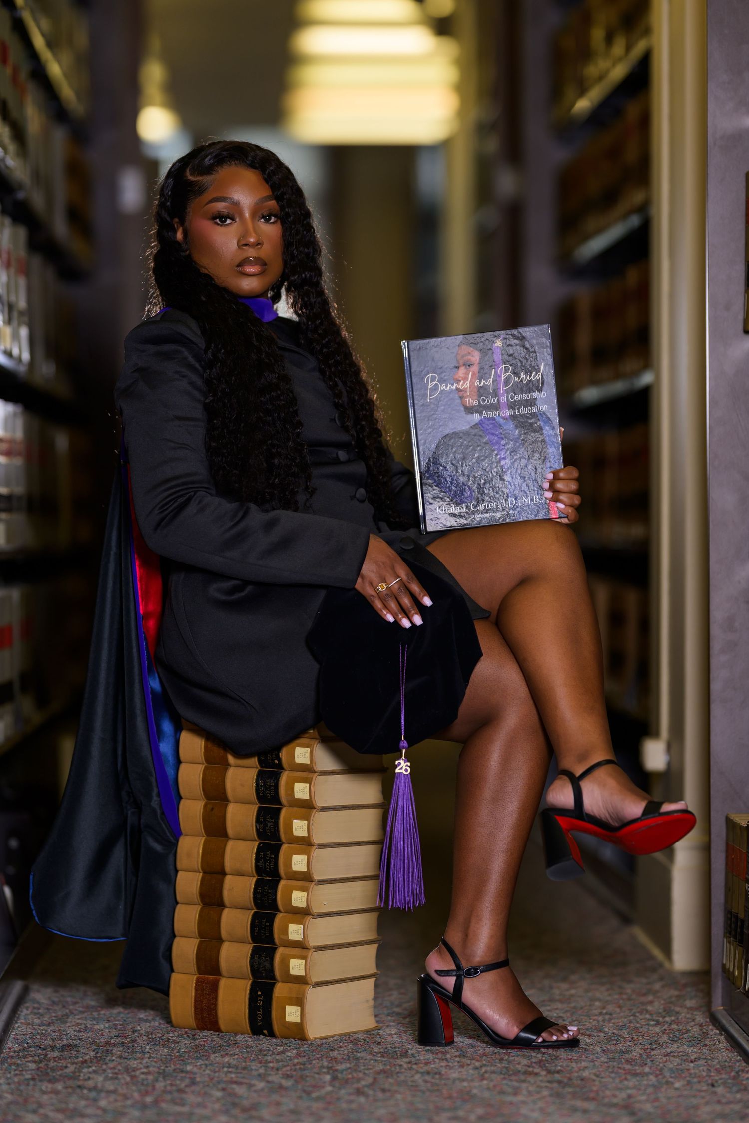 Woman in graduation attire holds a book titled Beautiful Proud while seated on stacked books in a library.
