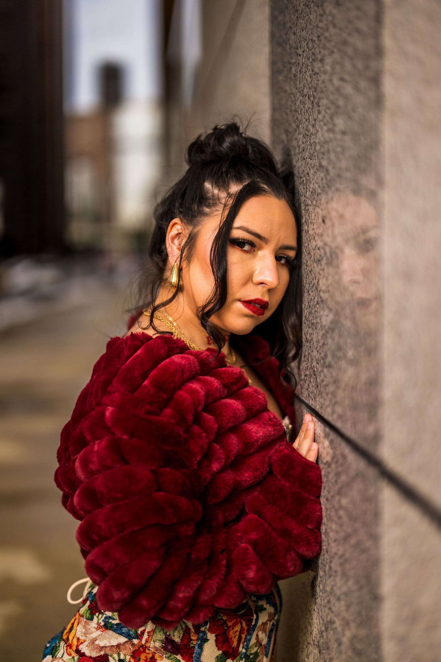 Woman in red faux fur jacket leans against concrete wall in urban alley, wearing floral dress and gold earrings.