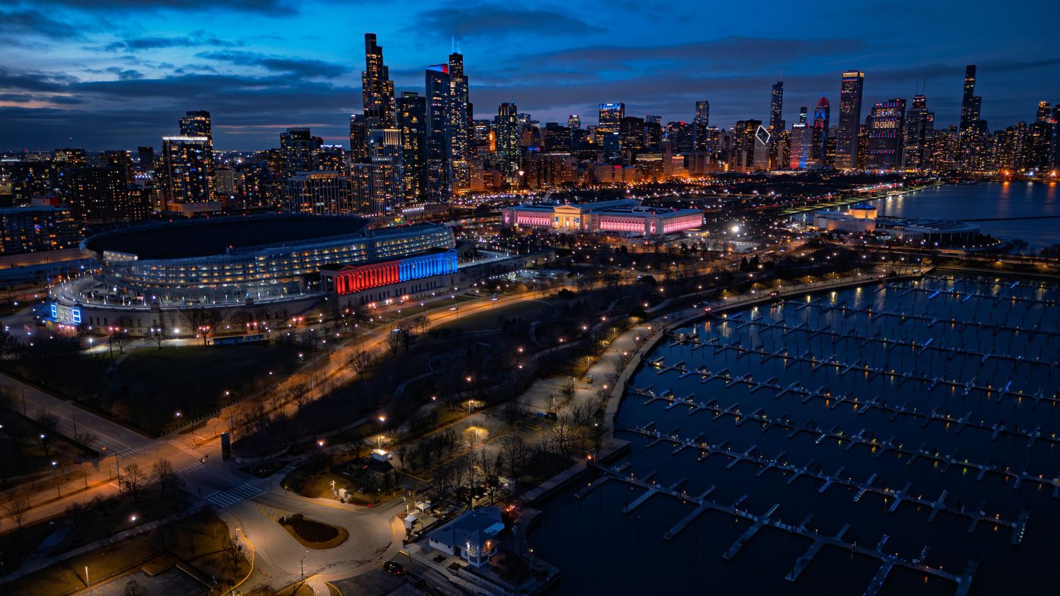 Aerial night view of Chicago skyline with illuminated skyscrapers, lakefront marina, and colorful arena lights at dusk.