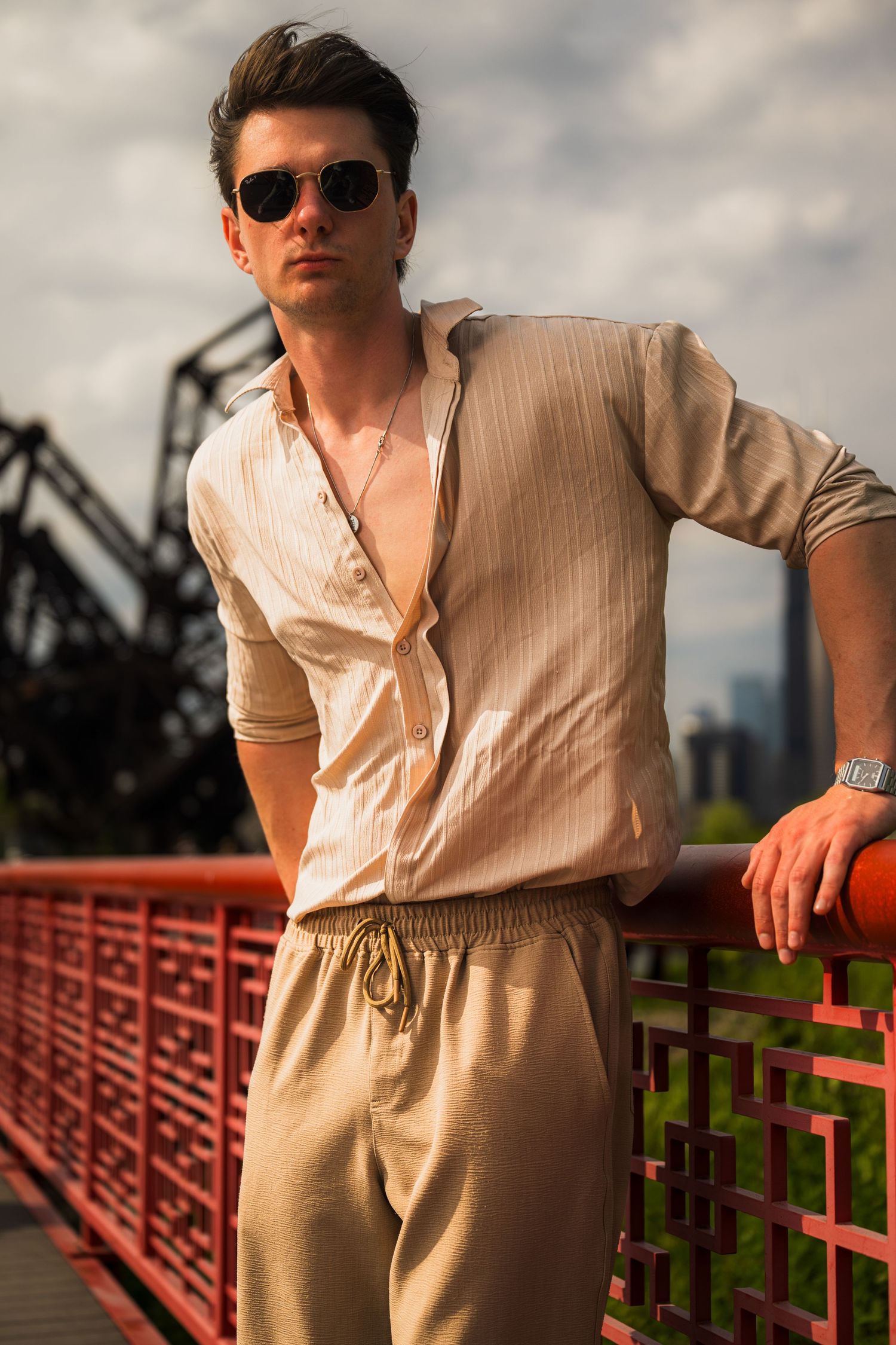 Stylish man in beige linen outfit and sunglasses leaning on red bridge railing with city skyline backdrop.