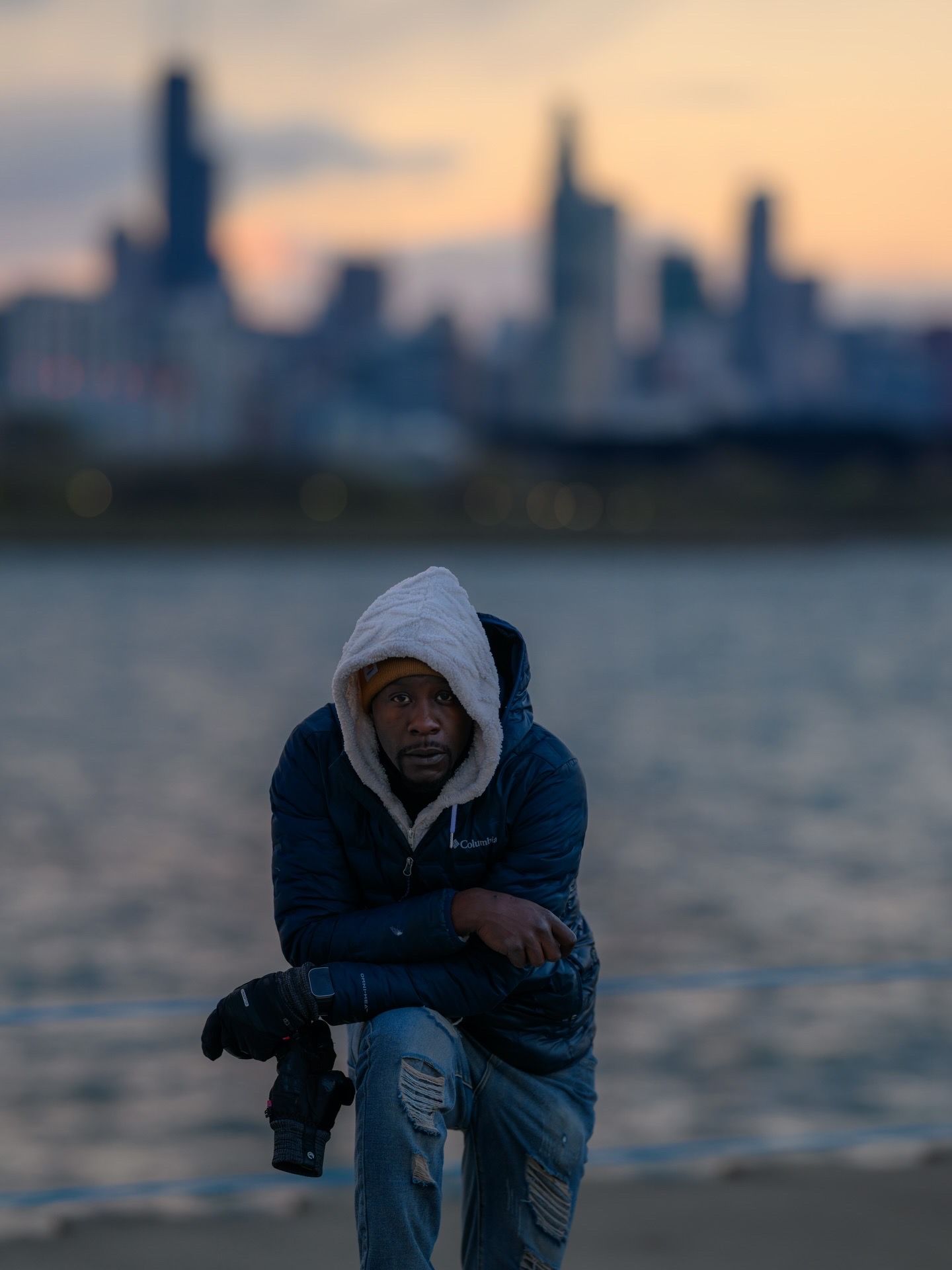 Man in hoodie sits by lakefront at dusk with Chicago skyline glowing behind him, holding a camera.