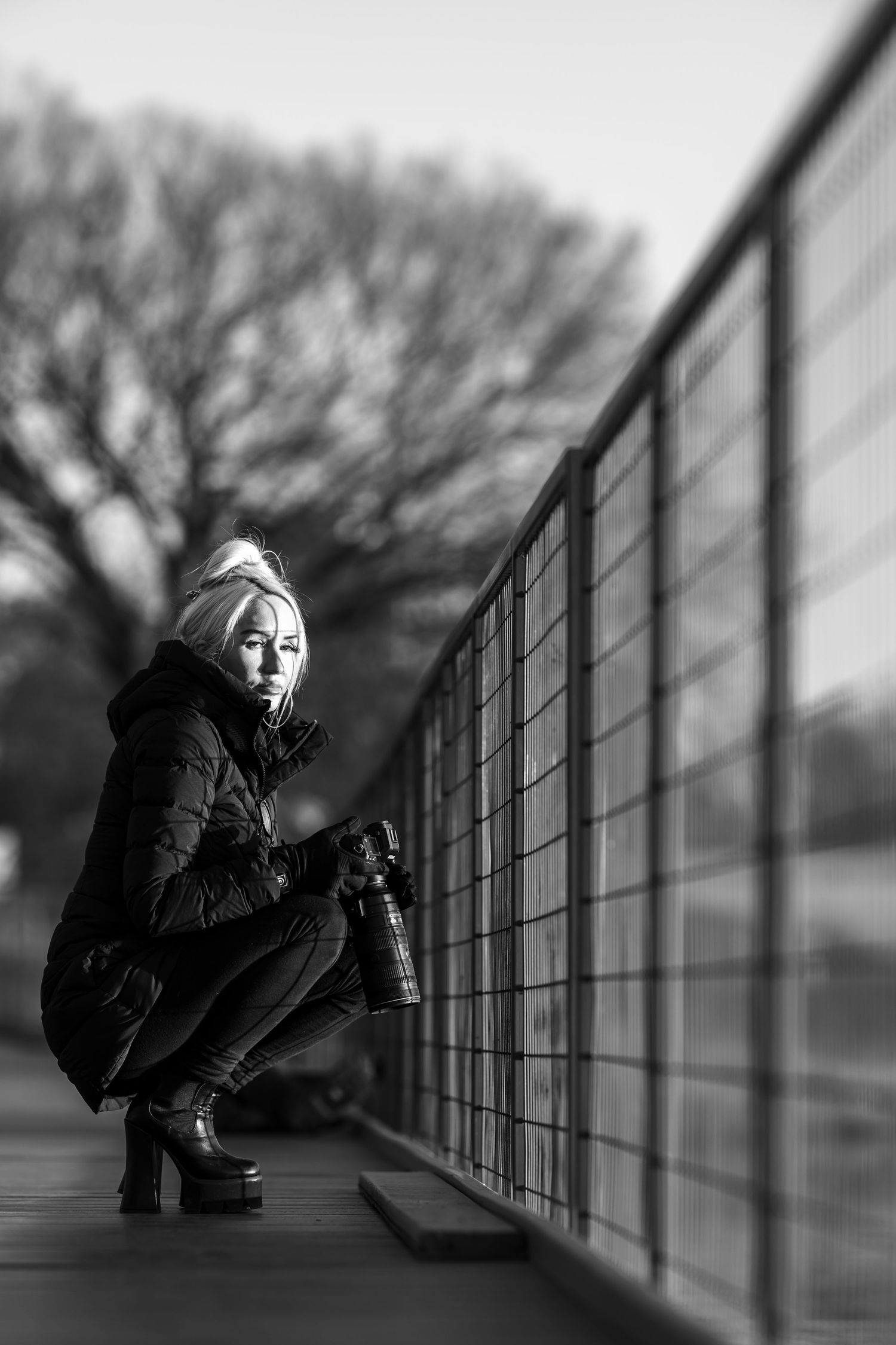Black and white photo of a blonde woman crouching by a metal fence outdoors, holding a camera.