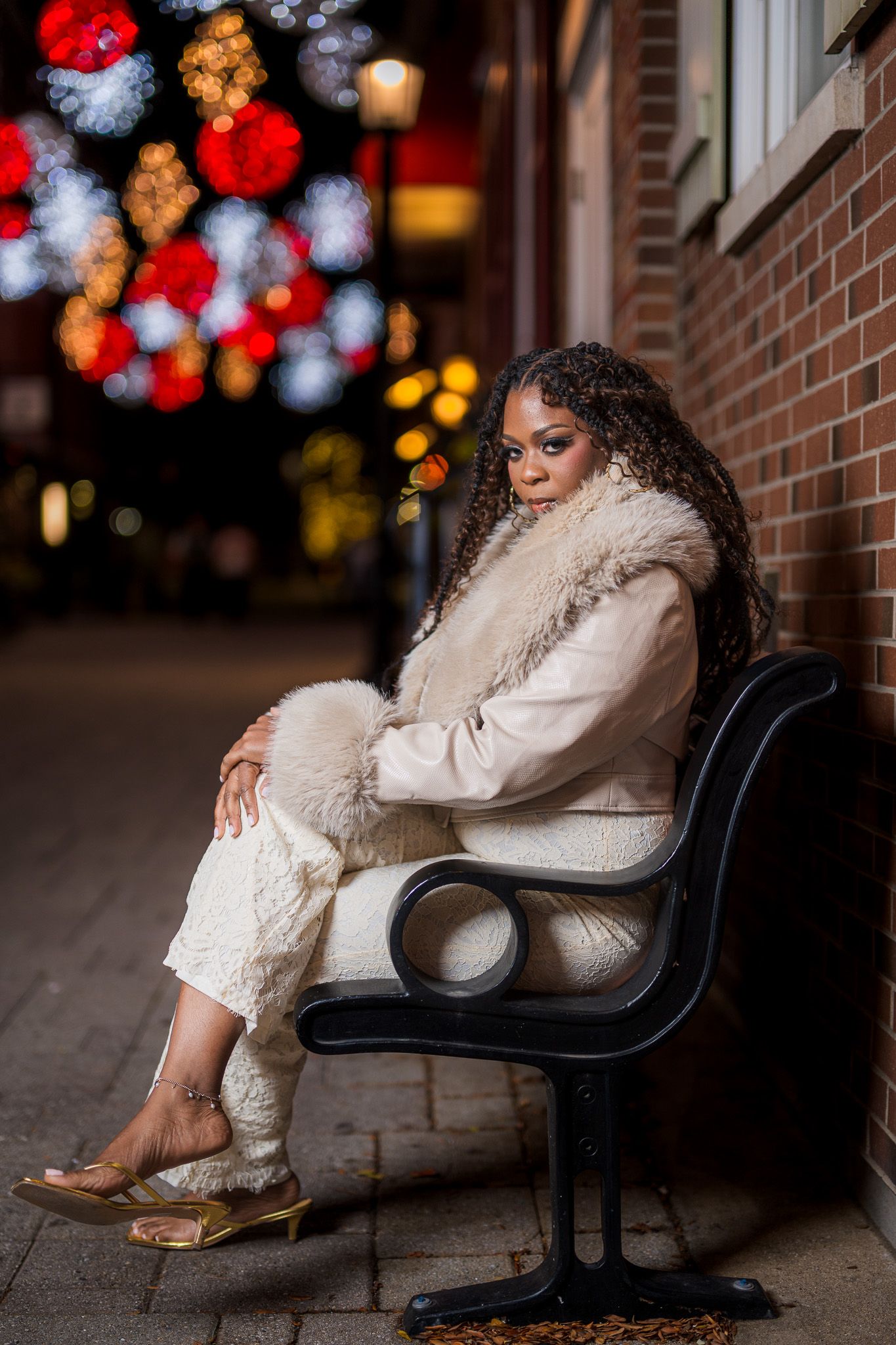 Woman in cream fur coat sits on bench at night with colorful bokeh lights behind her.