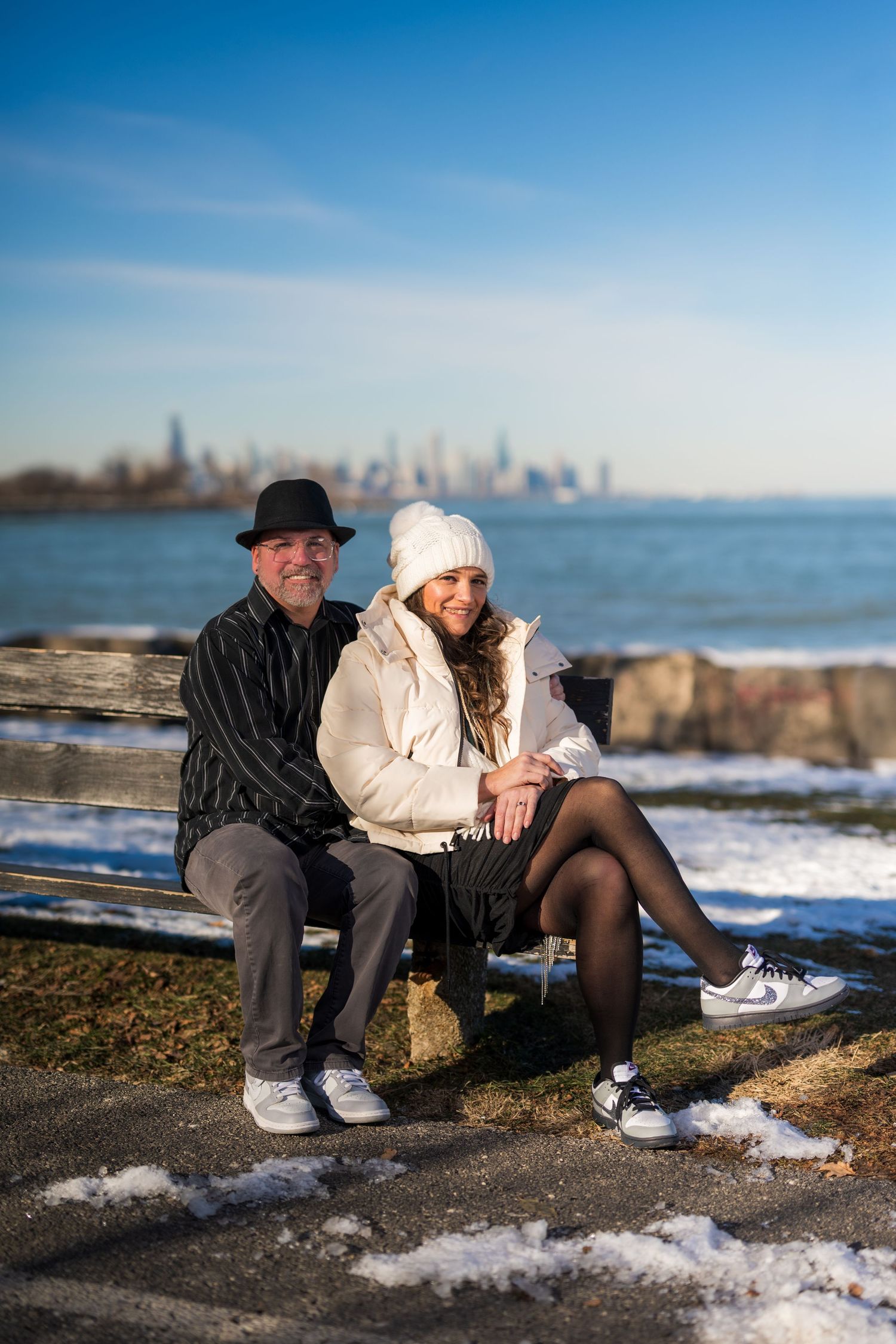 Couple sitting on a bench by the lakeside with a city skyline in the background on a sunny winter day.