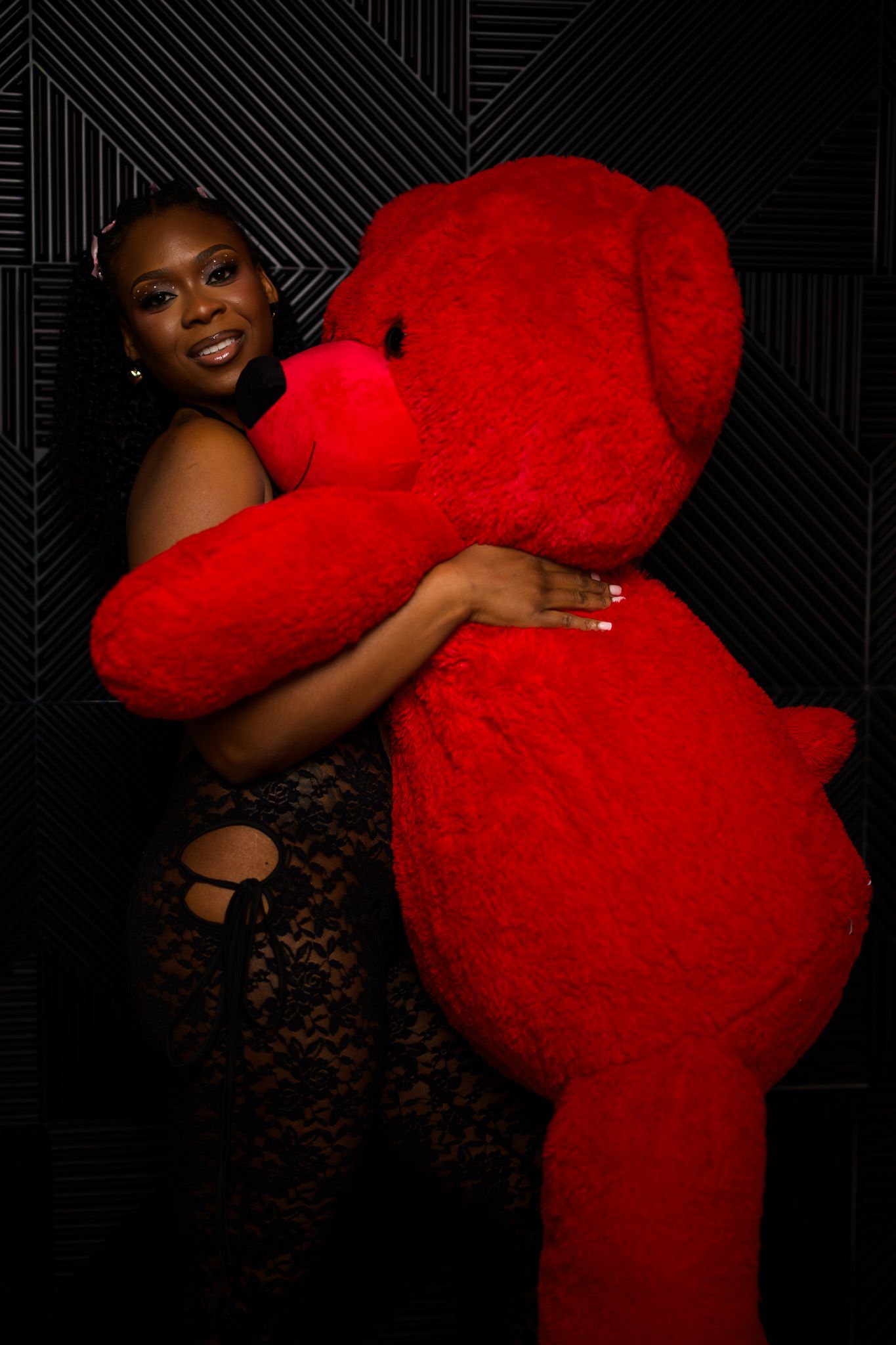 A smiling woman in a red dress joyfully hugs a giant red teddy bear against a dark geometric background.