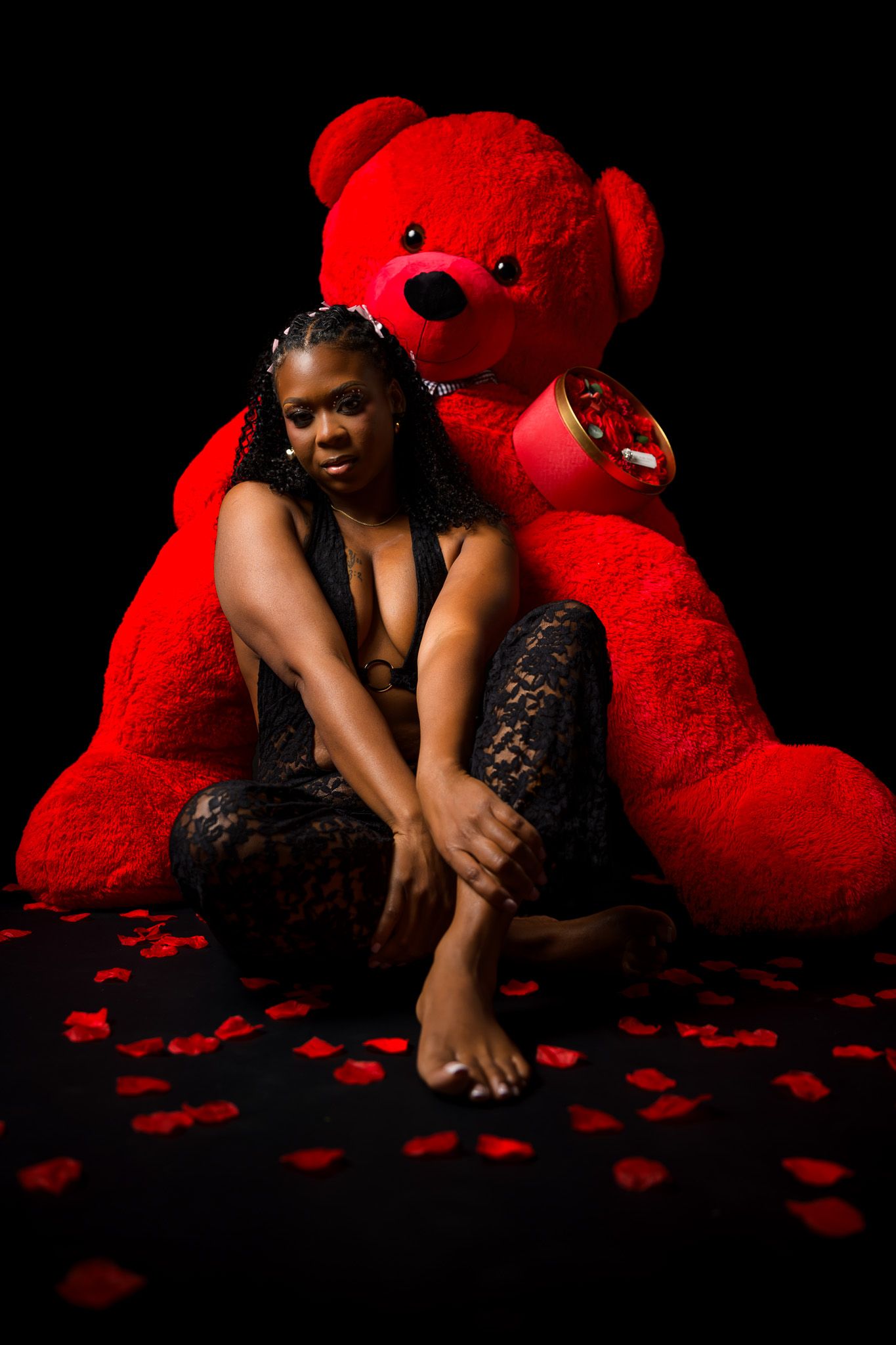 Woman in black lace outfit poses with large red teddy bear surrounded by rose petals on dark background.