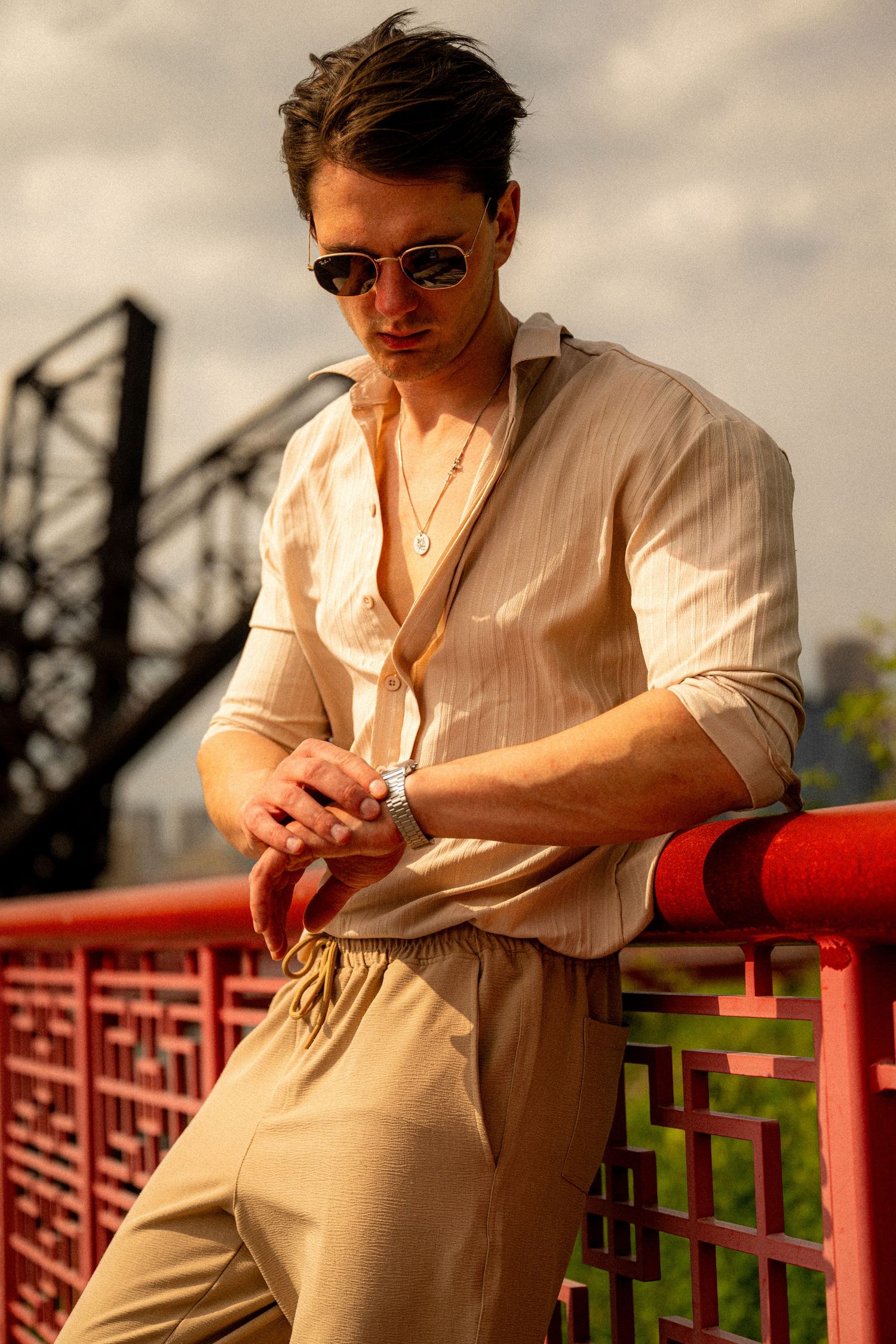 Stylish young man in beige linen outfit and sunglasses leaning on red railing outdoors.
