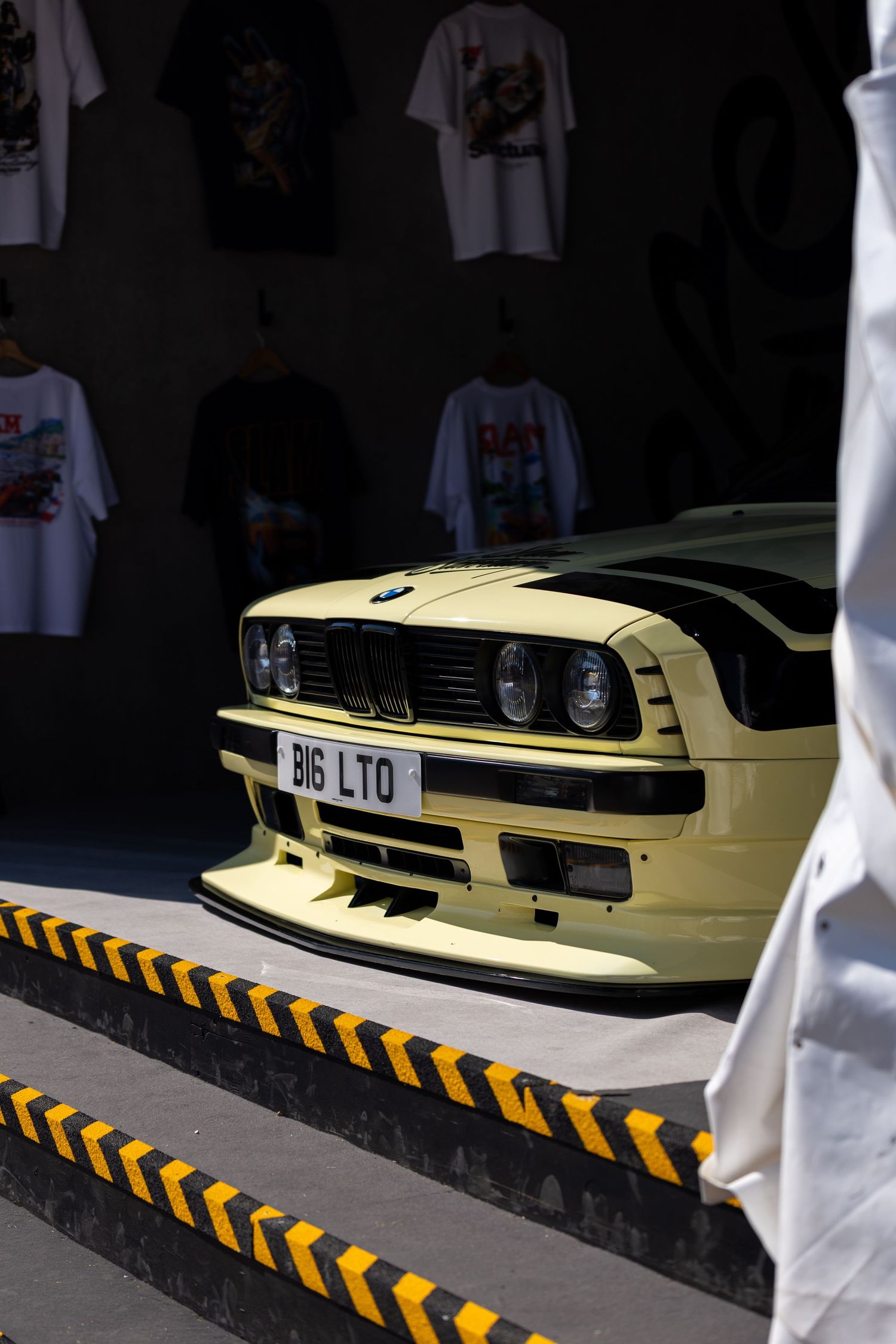 Yellow BMW E30 M3 with BIG LTD plate on display, framed by caution tape and t-shirts in background.