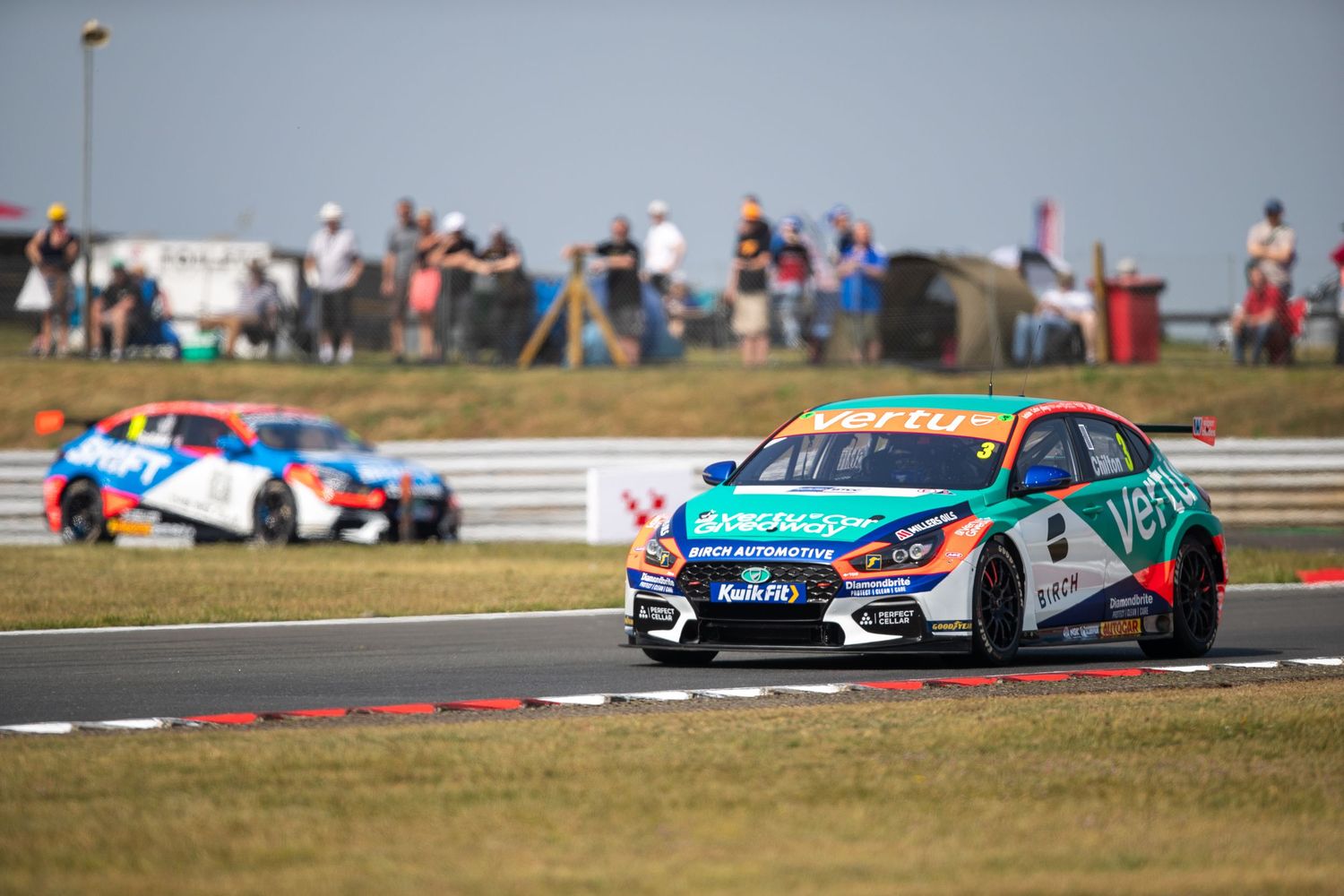 Two colorful touring cars race on a circuit track with spectators watching from the hillside on a sunny day.