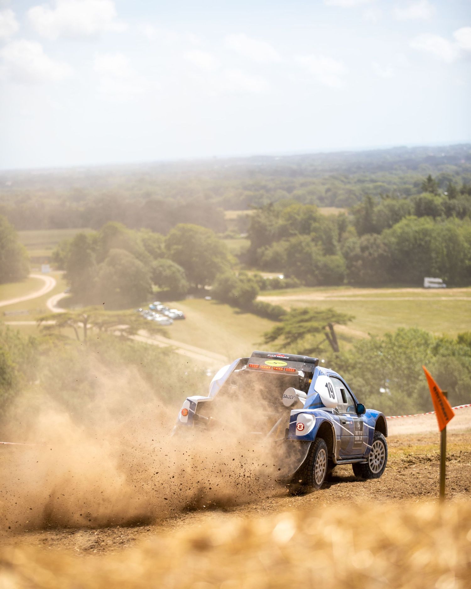 Rally car kicking up dust on a dirt stage with scenic countryside landscape behind.