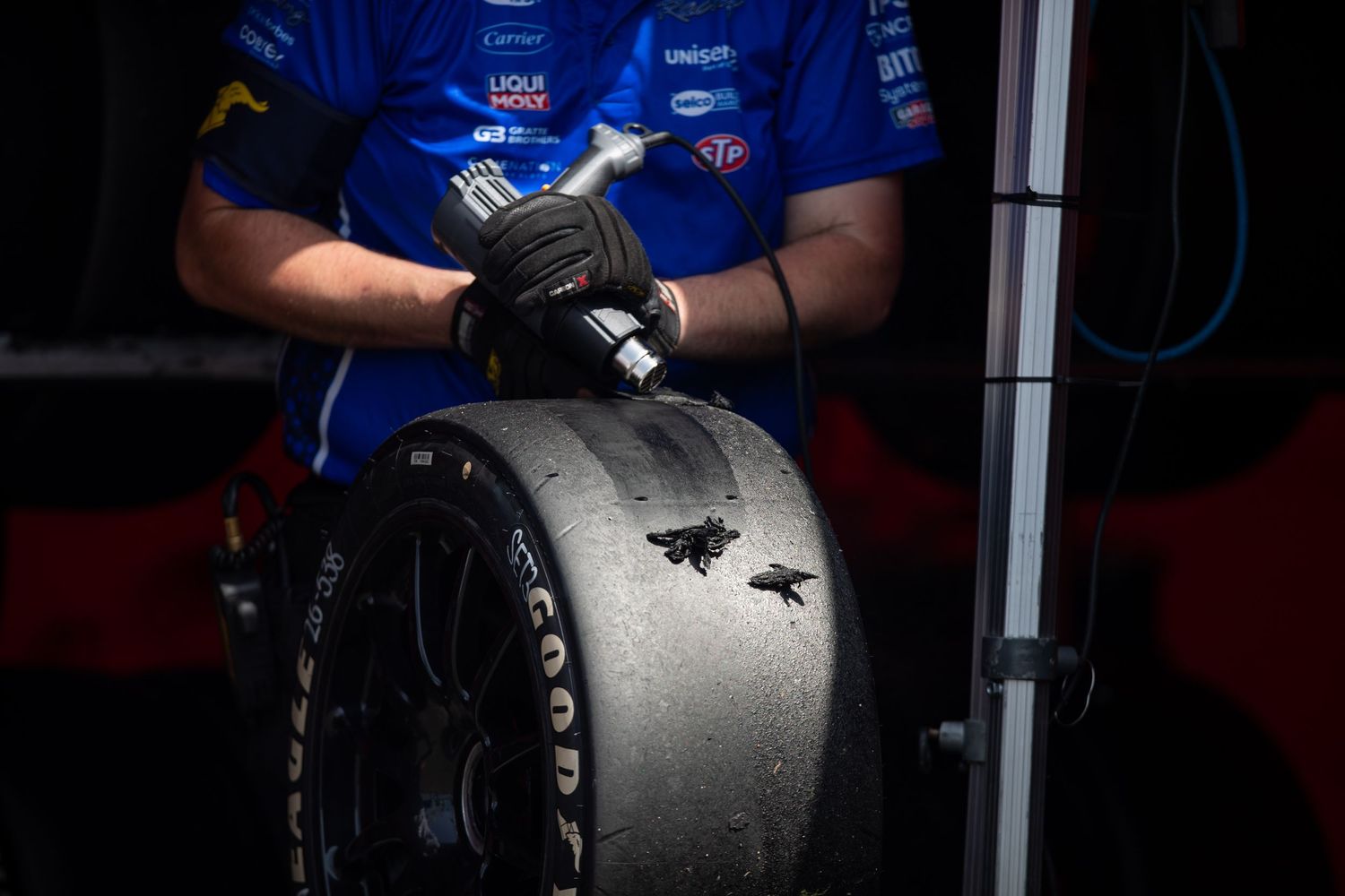 Racing mechanic in blue uniform handling a worn racing slick tire in the pit garage during a motorsport event.