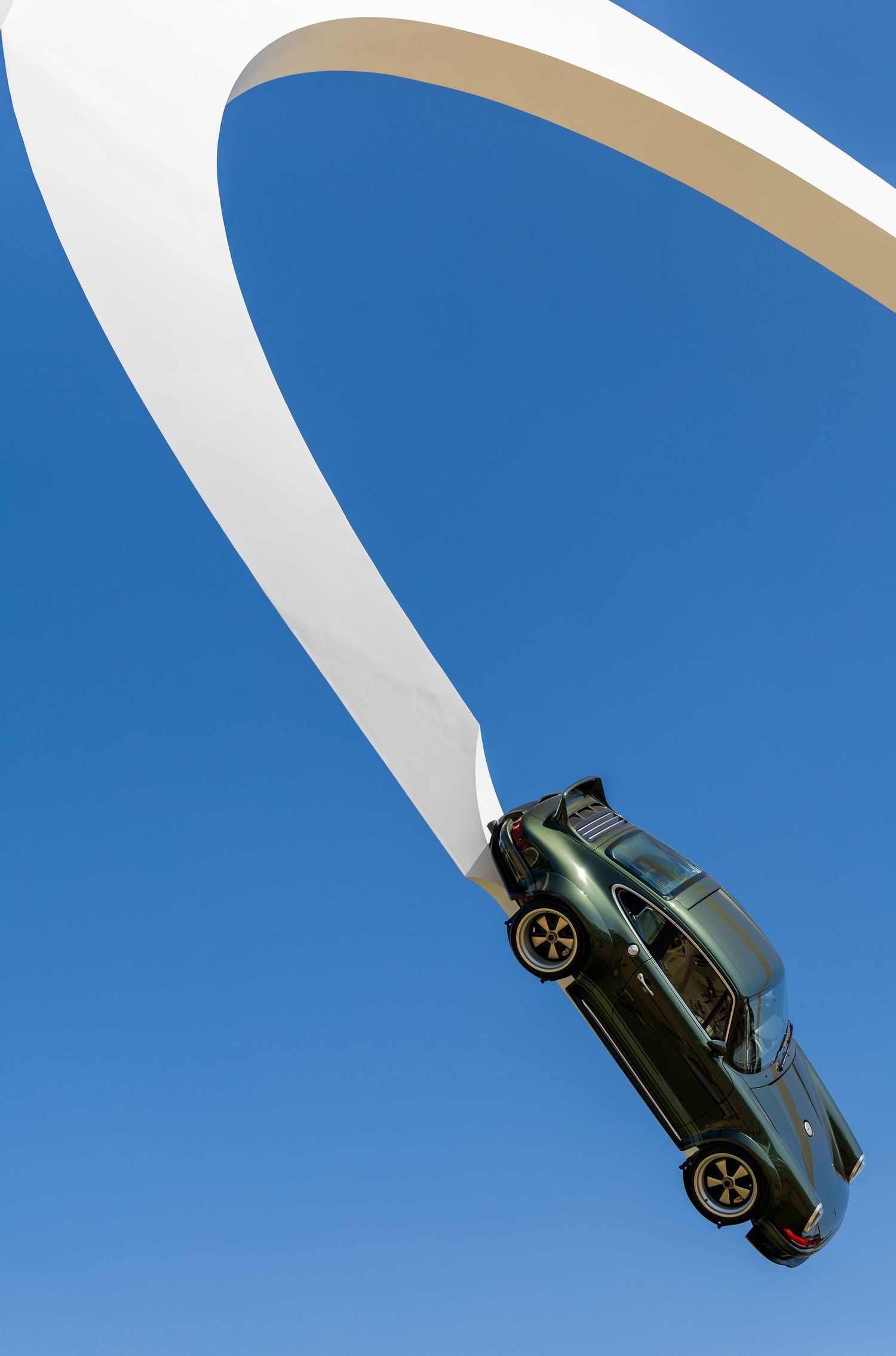 A dark green sports car mounted vertically on a sweeping white arch sculpture against a clear blue sky.