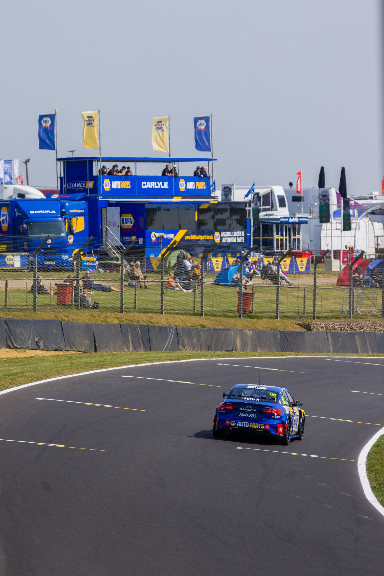Blue BTCC race car on circuit with NAPA branded pit lane infrastructure in background.