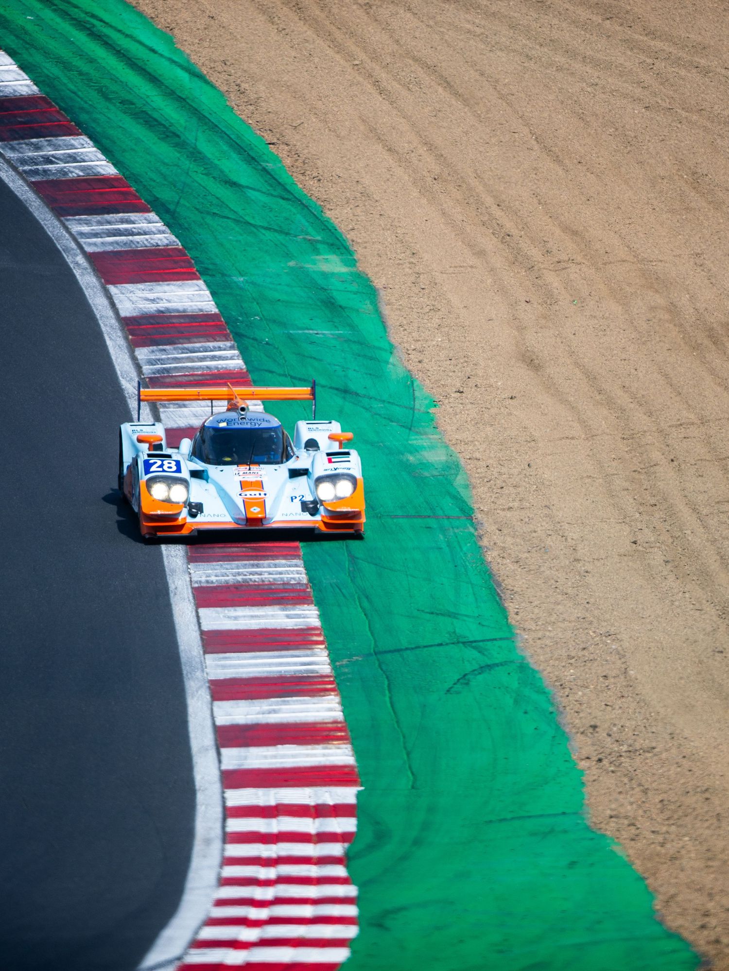 Gulf-liveried LMP race car #28 navigating a corner at a motorsport circuit with red and white curbing.