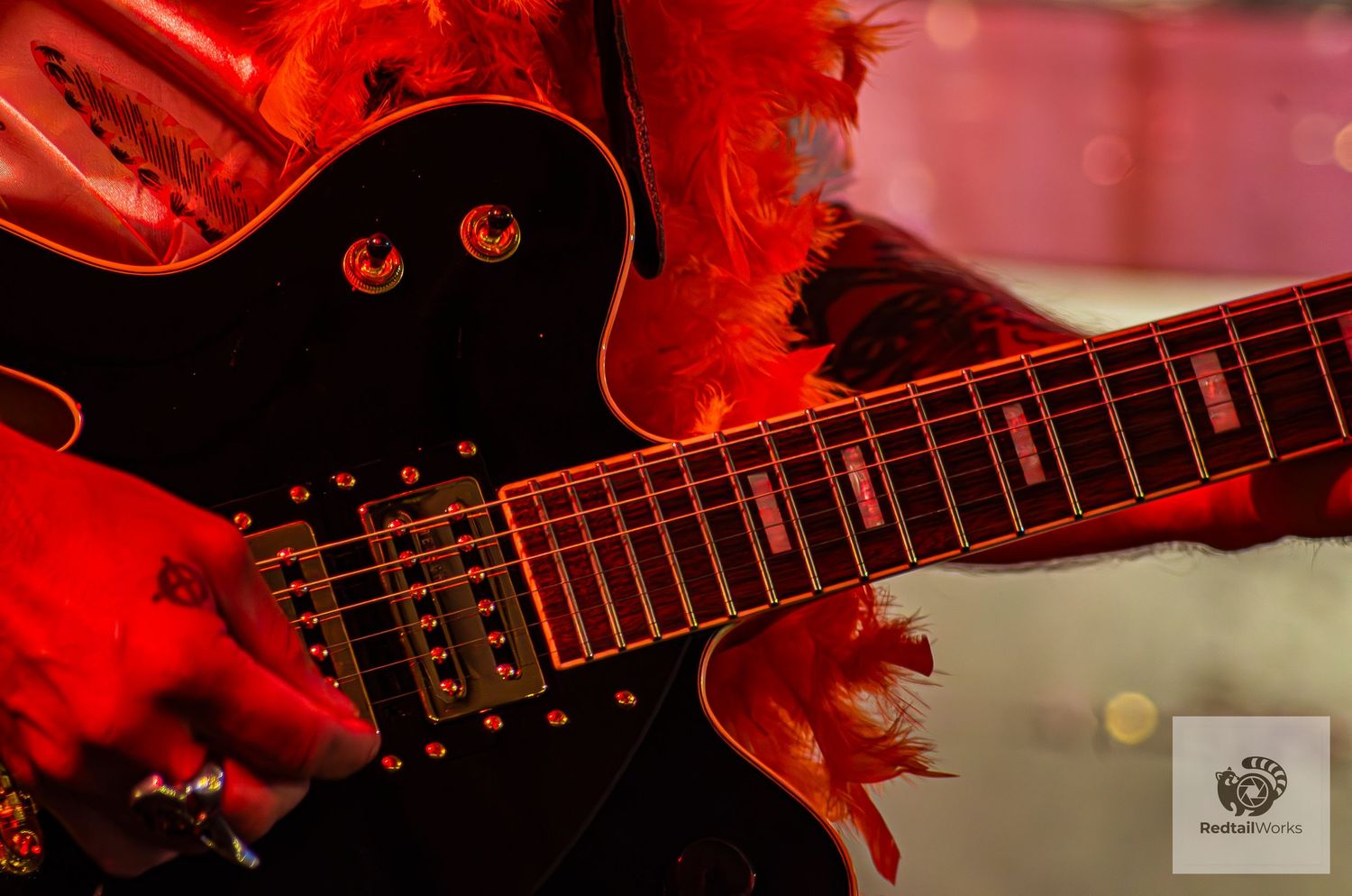 Close-up of black electric guitar with red feathers and gold hardware under vivid red stage lighting.