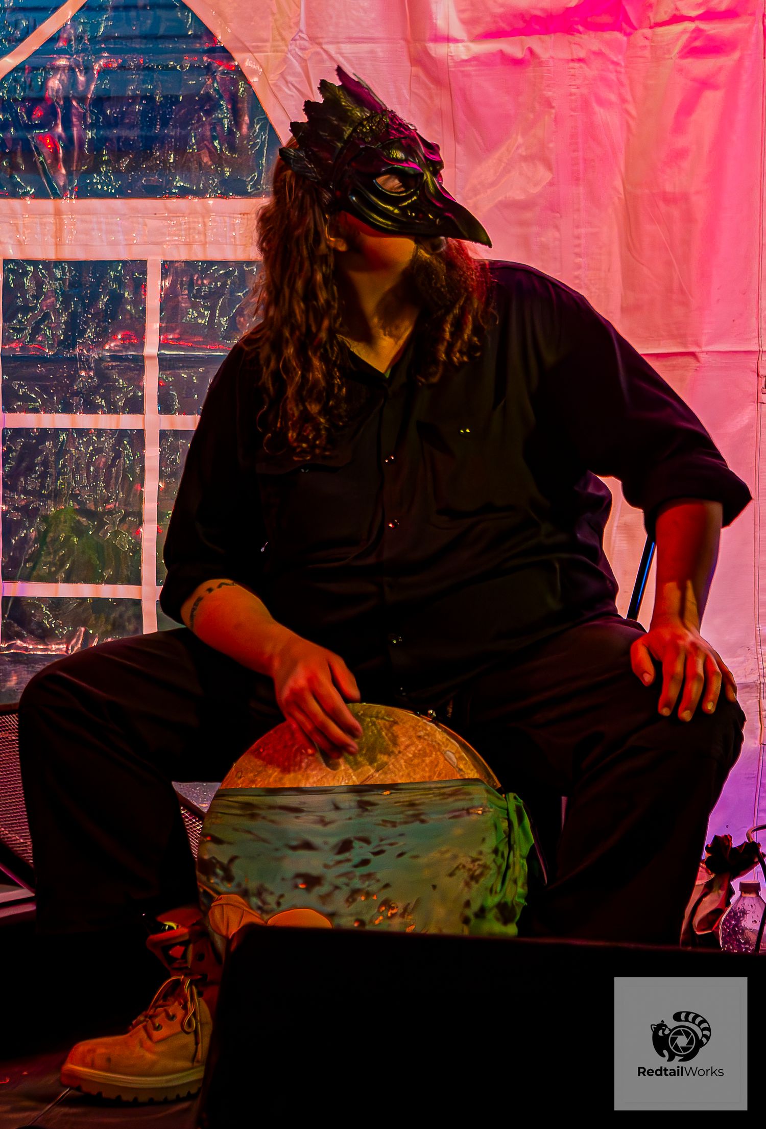 Masked performer in a bird mask playing a drum on a colorfully lit stage.