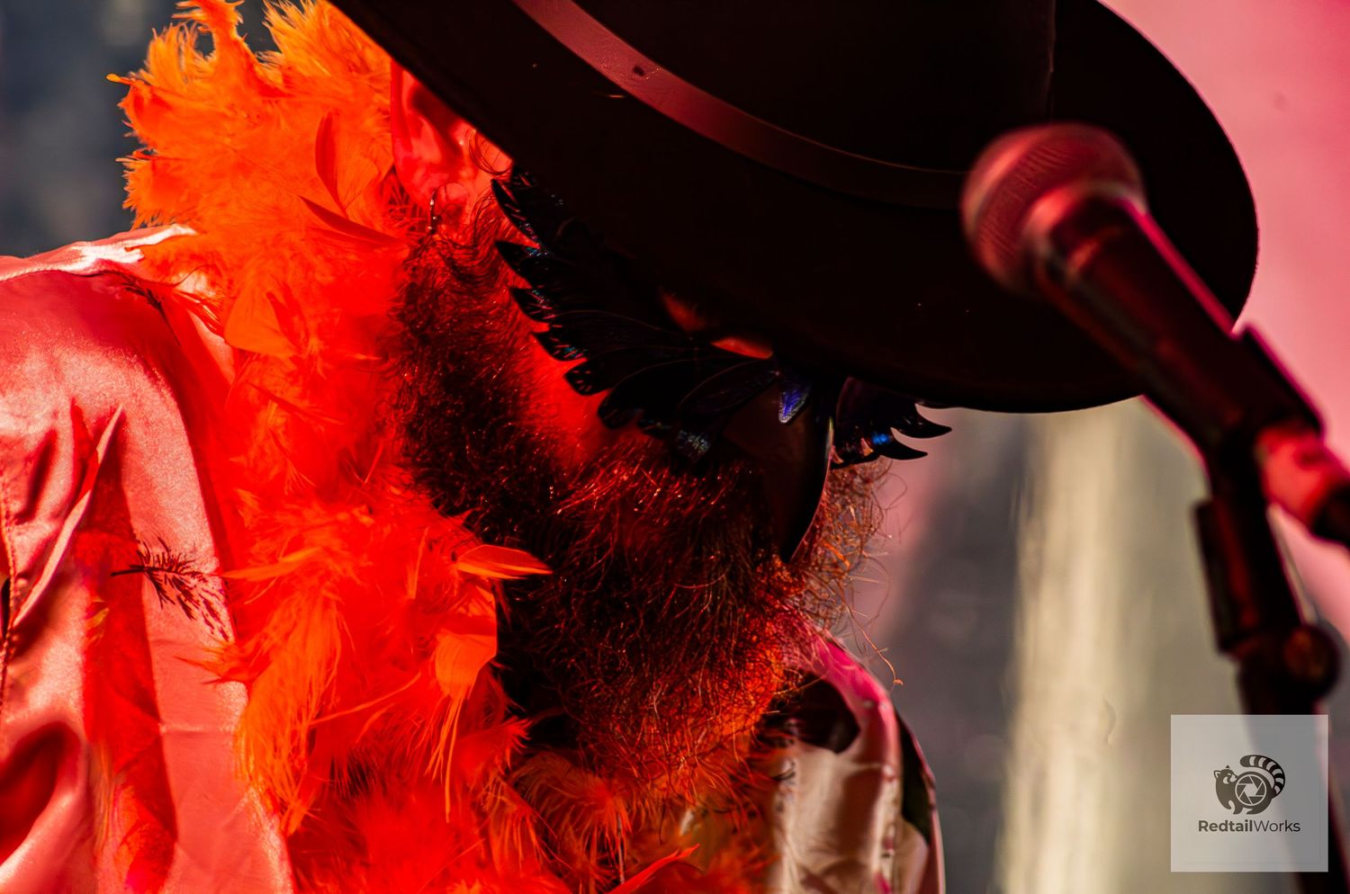 Musician wearing black hat and vibrant orange feather boa performs on stage under red lighting.