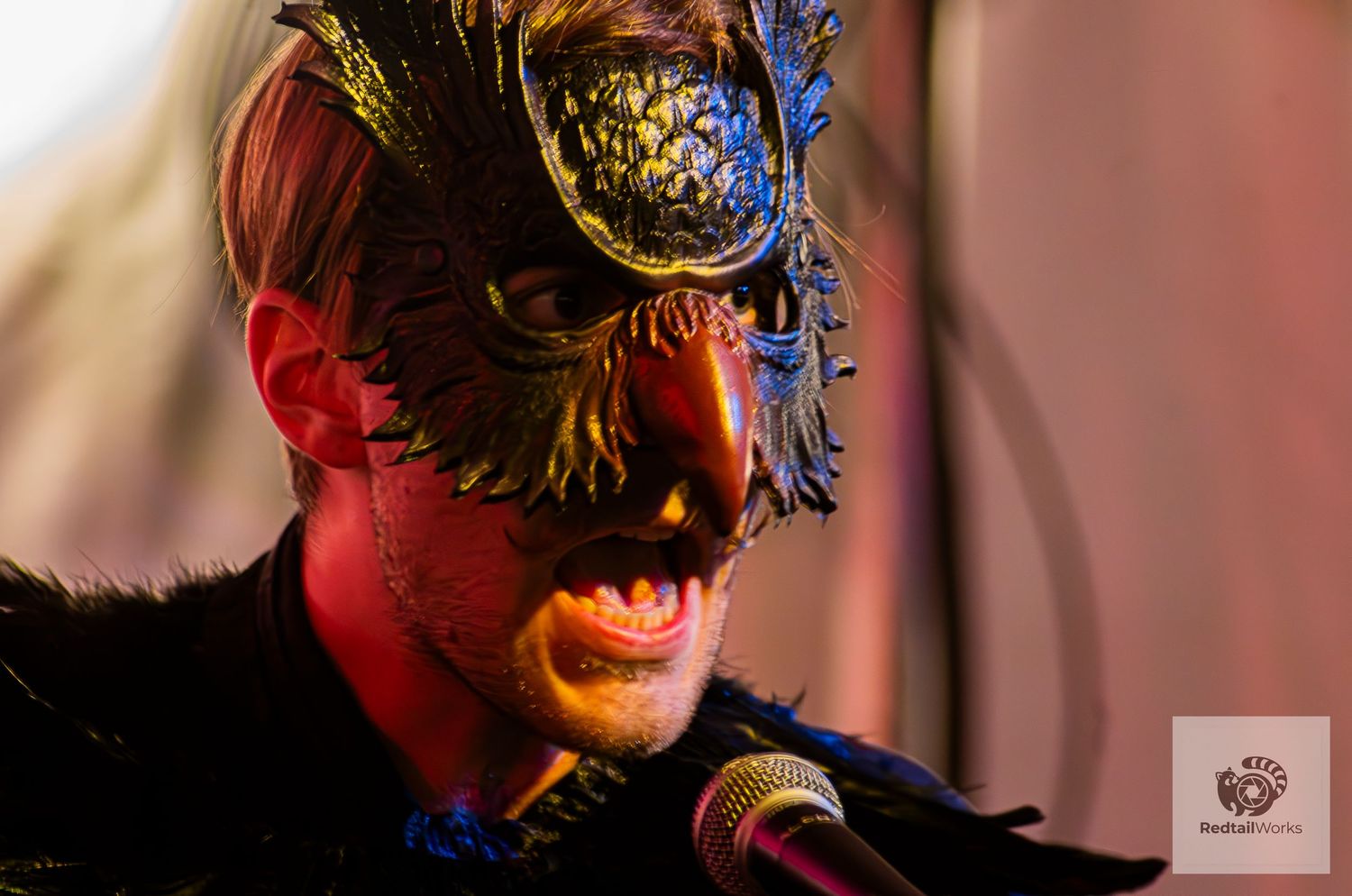 Performer wearing ornate gold masquerade mask sings into microphone under dramatic red stage lighting.