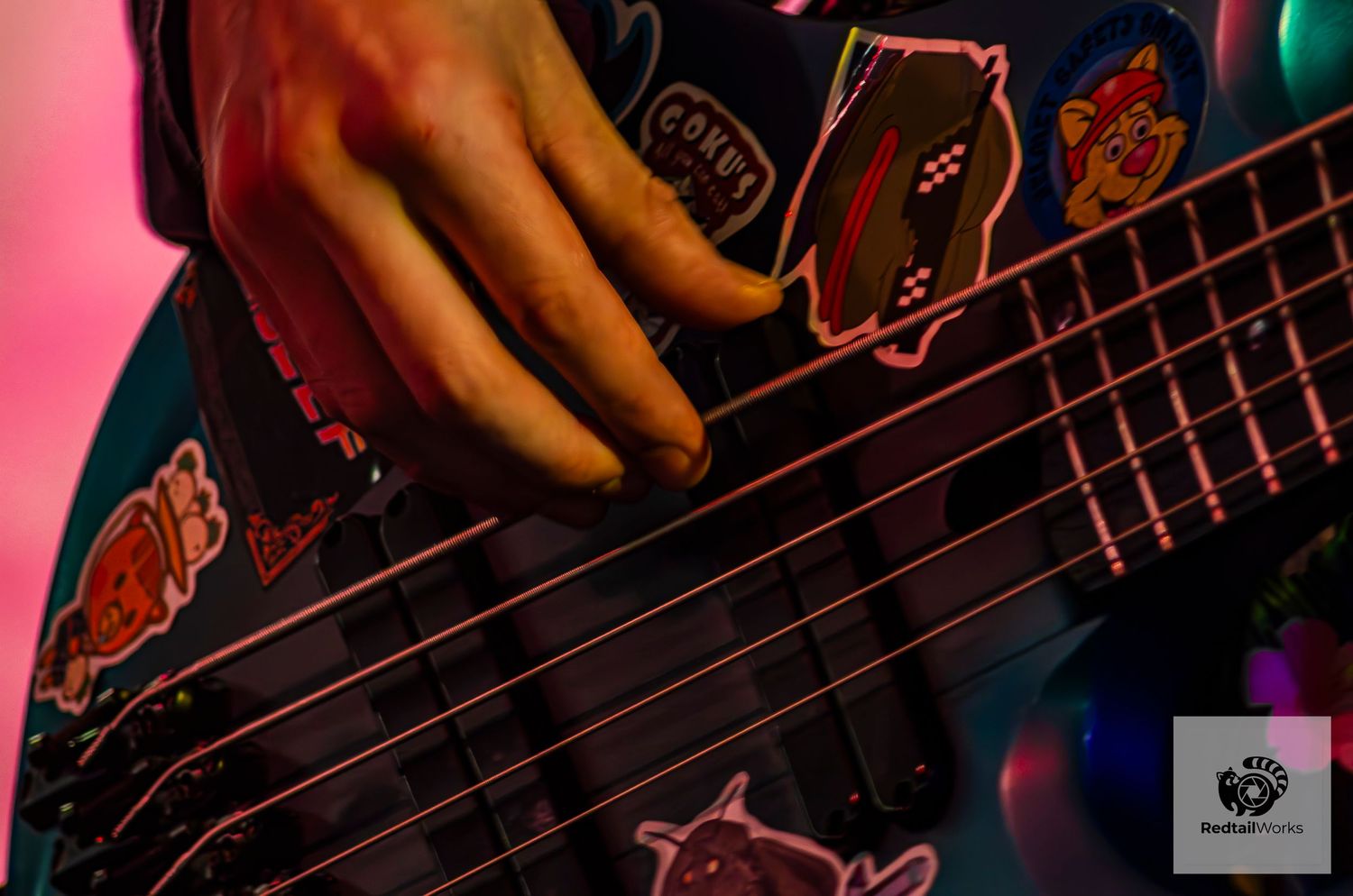 Close-up of musician's hand playing a decorated bass guitar with colorful stickers under pink stage lighting.