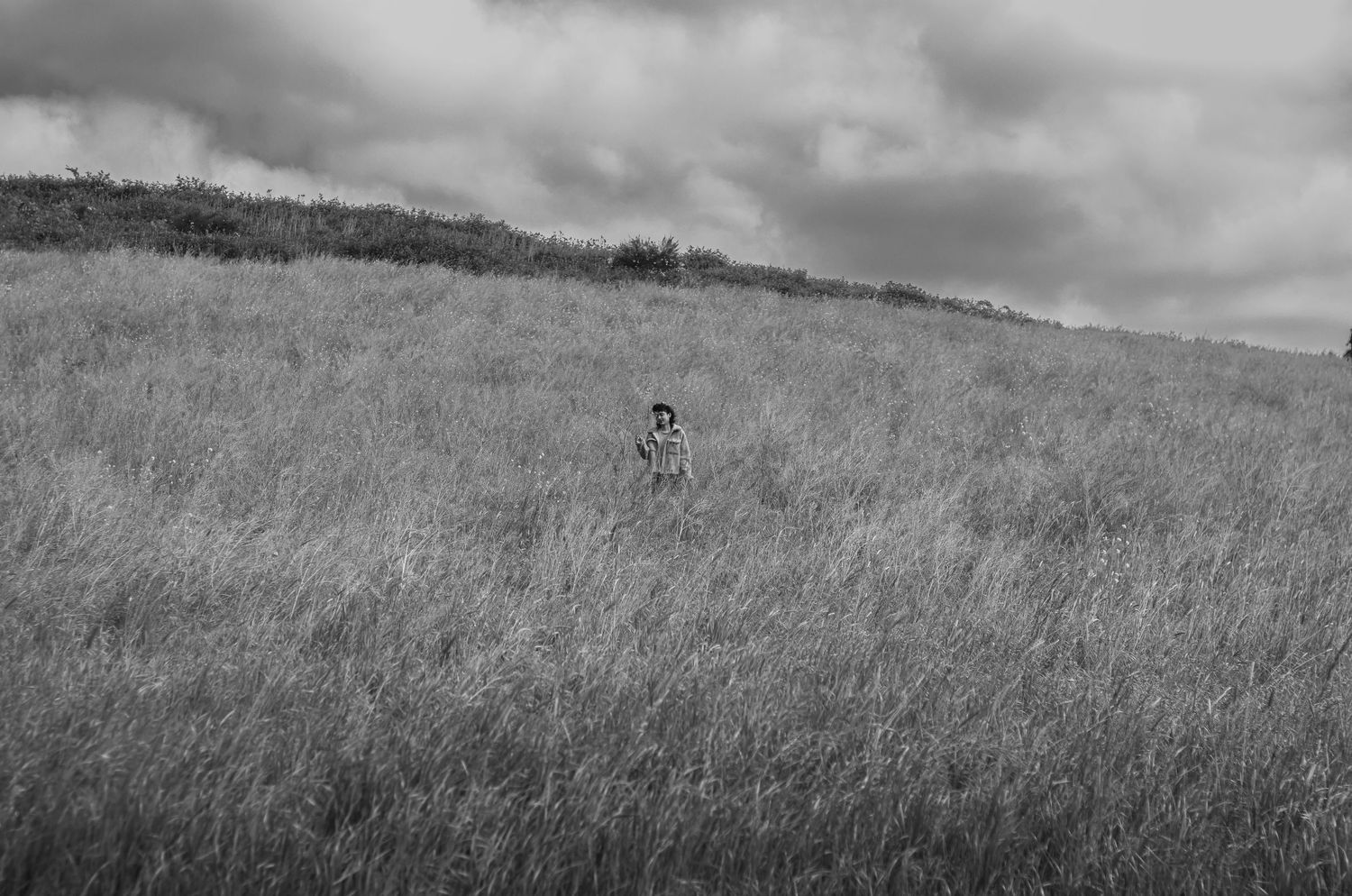 Black and white photo of lone person standing in tall grass field under dramatic cloudy sky.